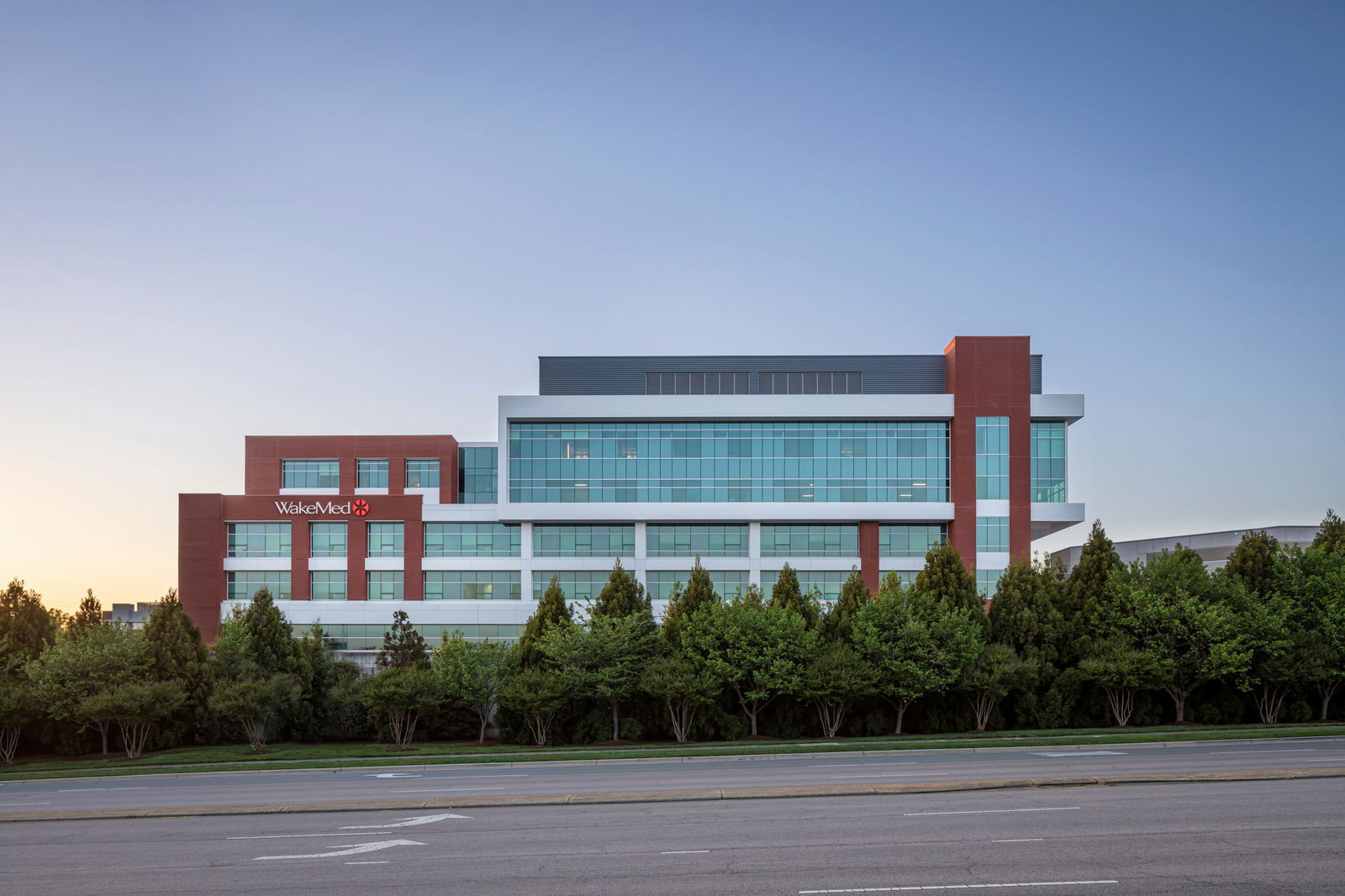 View of the WakeMed Cary Hospital, a modern medical facility building with red brick and light metal exterior with large glass windows from the highway. The building has a 'WakeMed' logo on the brick and is against a clear blue sky at dawn or dusk.