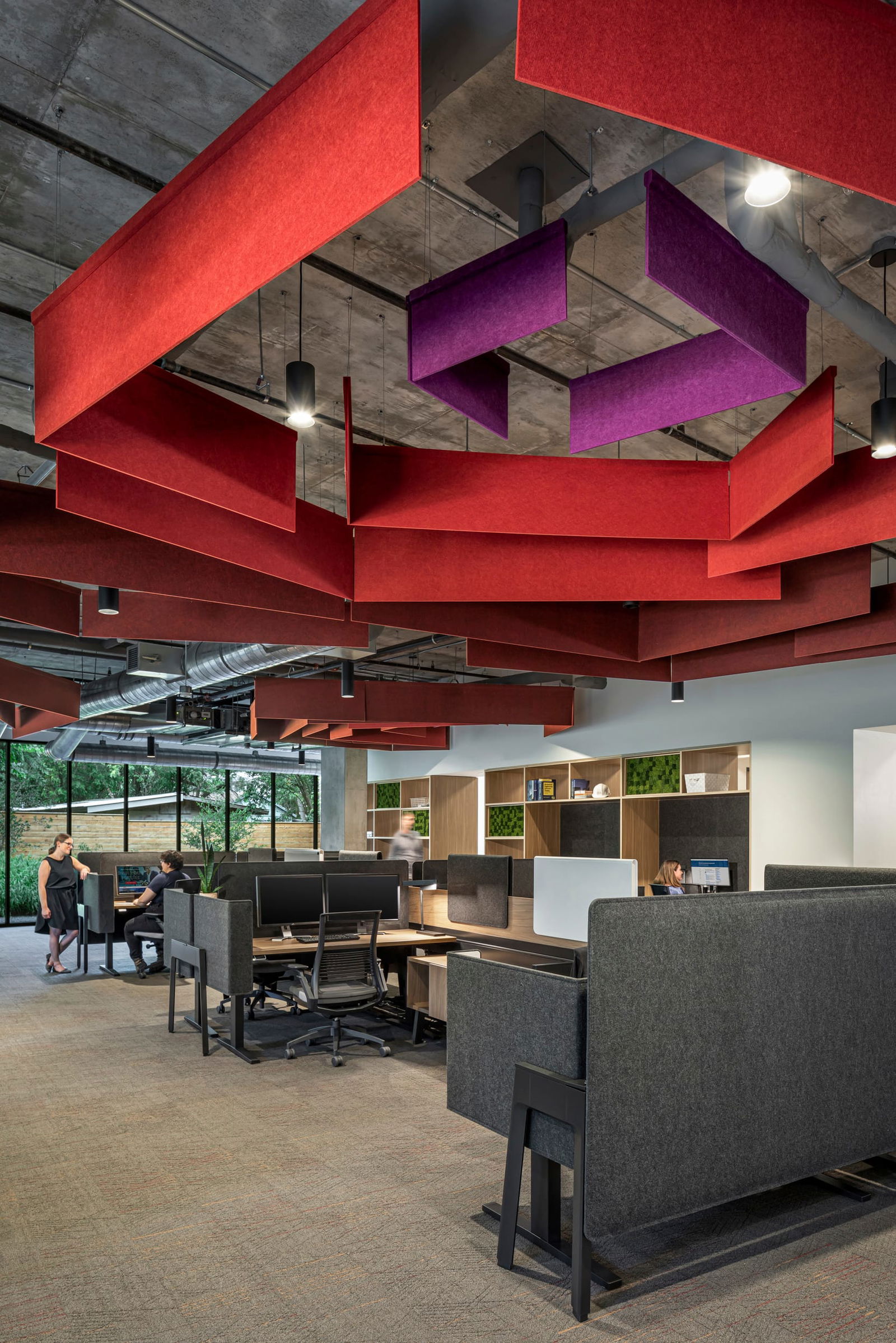 This image showcases BSA's open office workspace featuring distinctive acoustic ceiling elements with bold red baffles arranged in geometric patterns, complemented by purple accent panels. The space incorporates semi-private workstations with charcoal gray acoustic dividers creating individual zones while maintaining an open environment. Workers are visible at ergonomic desks with multiple monitors. Natural light floods in through large windows, while the background displays BSA's signature wood shelving units with integrated green moss panels. The design balances functionality and aesthetics with exposed concrete ceiling structure, modern lighting, and a thoughtful mix of materials that demonstrates BSA's architectural expertise in creating collaborative yet focused work environments.