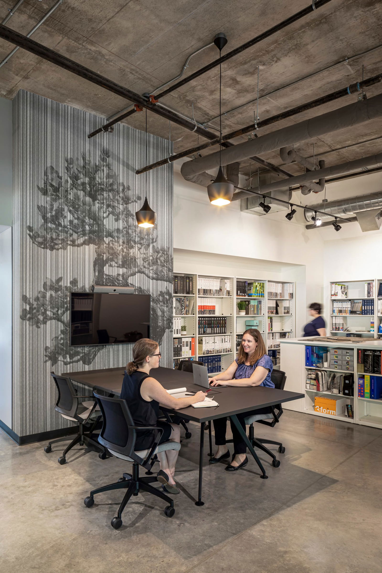 This image showcases a collaborative meeting space in BSA's office where two professionals engage in discussion at a dark wood conference table with ergonomic office chairs. The design features an artistic wall treatment with vertical striping and a subtle tree silhouette, creating visual interest while maintaining the sophisticated atmosphere. Pendant lighting with metallic finishes hangs from an exposed concrete ceiling that reveals the building's structural and mechanical systems. In the background, extensive white bookshelves house the firm's reference materials and resources, while polished concrete flooring completes the modern, industrial aesthetic. This thoughtfully designed meeting area exemplifies BSA's commitment to creating spaces that balance functionality with distinctive design elements that inspire creative collaboration.