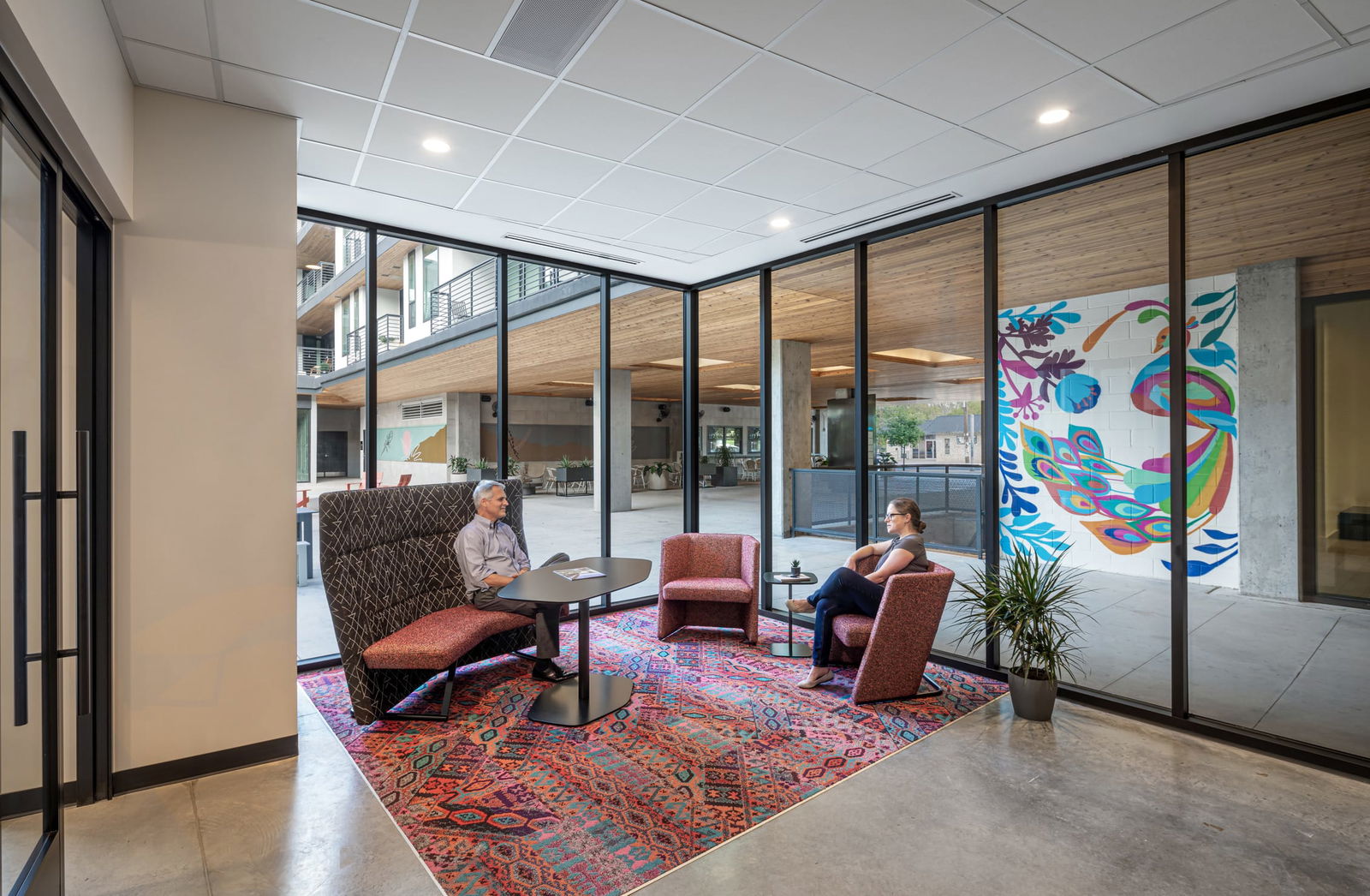 This image shows a modern meeting lounge in BSA's office featuring two professionals in conversation across a round black table. The seating area incorporates a high-backed patterned booth and complementary coral armchairs placed on a vibrant patterned area rug in reds and blues that adds warmth and color to the concrete flooring. Floor-to-ceiling glass walls create visual connectivity with an adjacent atrium space that features wooden ceiling elements, concrete columns, and a colorful mural visible in the background. The transparent design maintains openness while providing acoustic separation, demonstrating BSA's expertise in creating inviting, flexible meeting spaces that balance privacy with connection to the larger office environment.