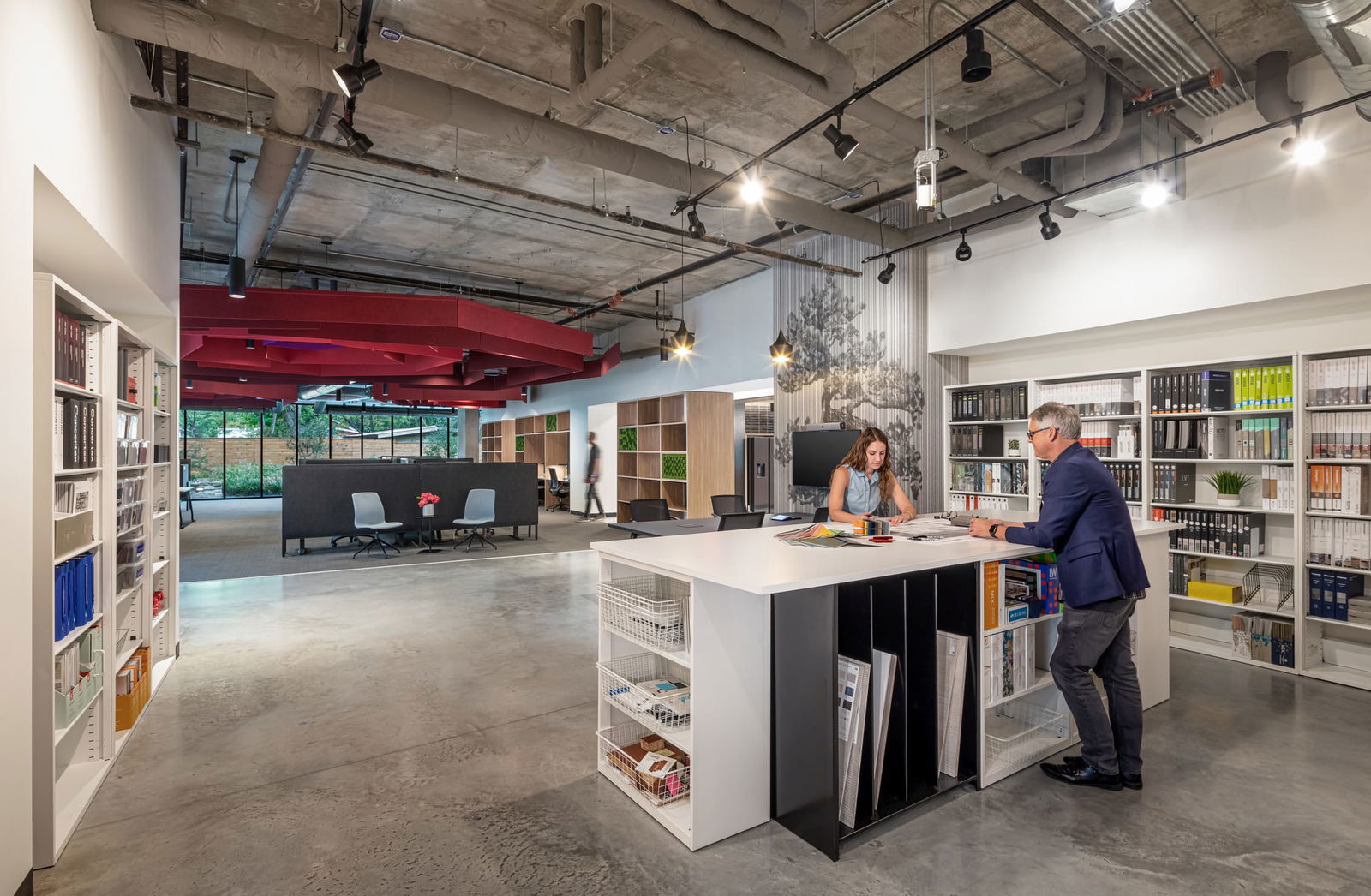 This image displays BSA's material library and collaborative workspace, where two professionals review material samples at a custom-designed island with integrated storage. The island features white countertops with black and white cubby storage below for material organization. Surrounding the space are extensive bookshelves housing material references, catalogs, and samples. The design maintains BSA's signature elements with red acoustic ceiling baffles, exposed concrete ceilings with mechanical systems, polished concrete floors, and light-filled work areas. A meeting area with black dividers and light-colored seating is visible in the background, while wood shelving units with green accents create semi-private zones. A decorative wall installation adds visual interest to this functional yet aesthetically sophisticated space that demonstrates BSA's design capabilities.