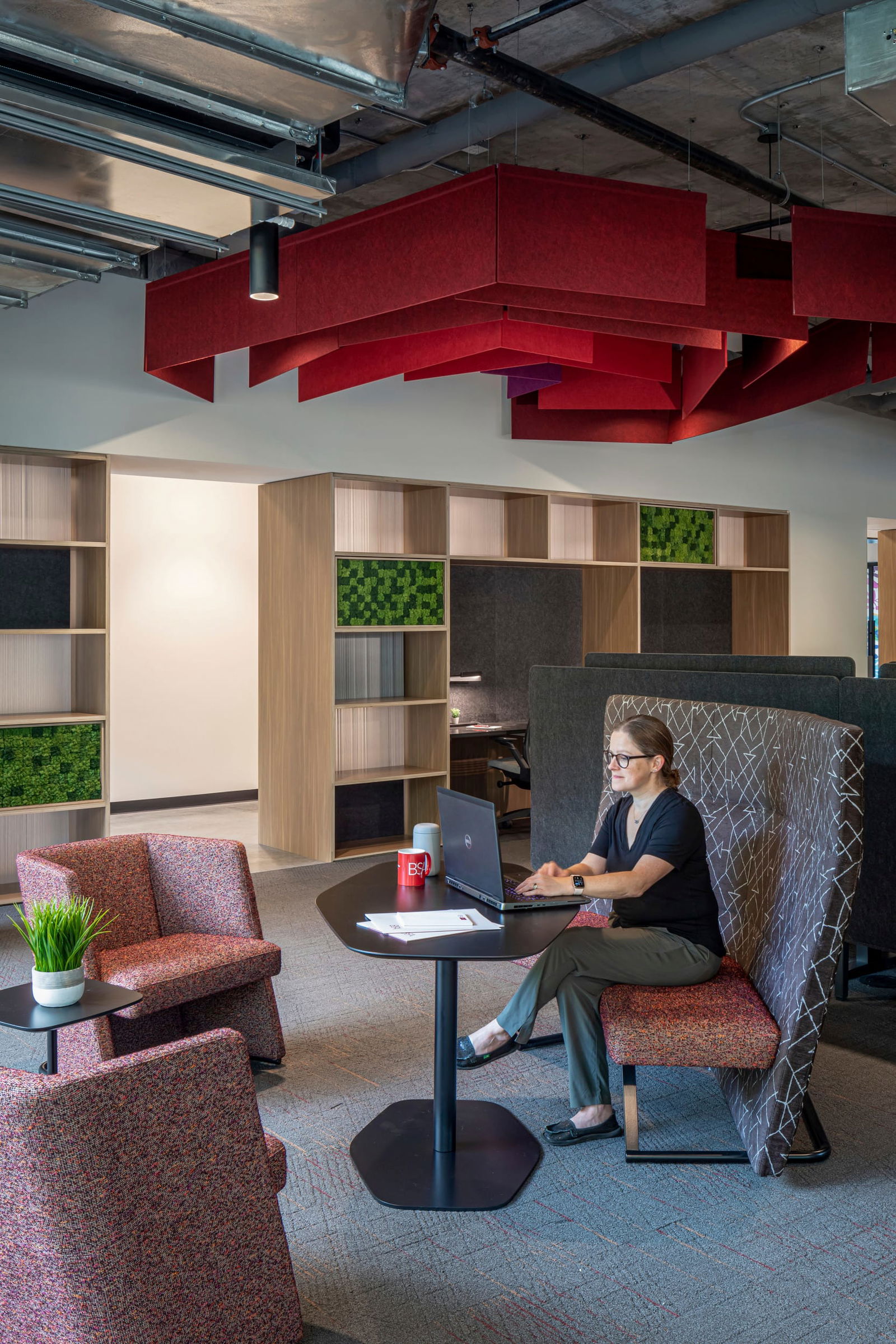 This image showcases an innovative collaborative workspace in BSA's office featuring distinctive red acoustic ceiling baffles suspended from an exposed concrete ceiling. A person works at a small black table with a laptop and a BSA-branded mug, seated in a high-backed booth with geometric patterned upholstery. The seating area includes textured red armchairs and small accent tables with plants. Behind the workspace, custom-designed wood shelving units with integrated green moss panels and acoustic elements create a semi-private environment while maintaining an open feel. The thoughtful interior design balances acoustics, privacy, and aesthetics with a warm material palette, highlighting BSA's expertise in creating functional yet visually striking professional environments.