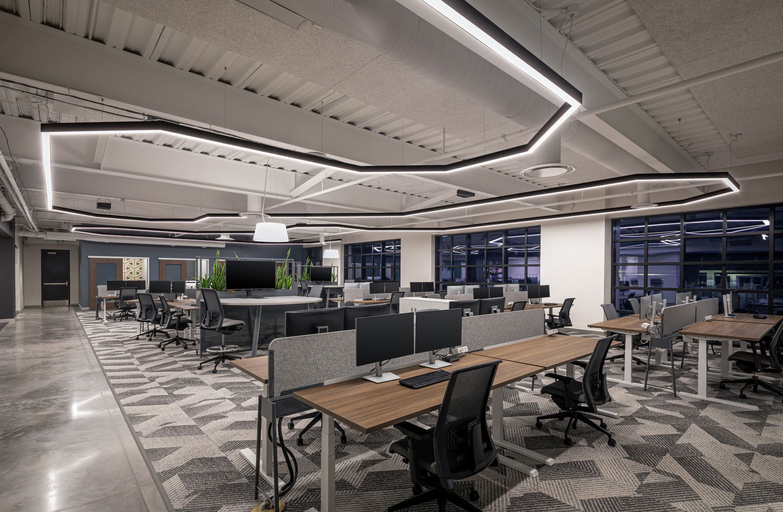 This image captures a sophisticated, high-performance workspace designed by BSA, showcasing a seamless blend of modern industrial and biophilic elements. The open office features clean-lined workstations with warm wood surfaces and acoustic privacy panels, all arranged on a dynamic, patterned carpet. Overhead, sculptural LED lighting echoes the geometry of the space, while exposed ceilings and polished concrete floors enhance the contemporary aesthetic. Centralized collaborative hubs, enhanced by greenery and integrated technology, promote team engagement and flexibility. The design reflects BSA’s commitment to innovation, sustainability, and user-focused environments.