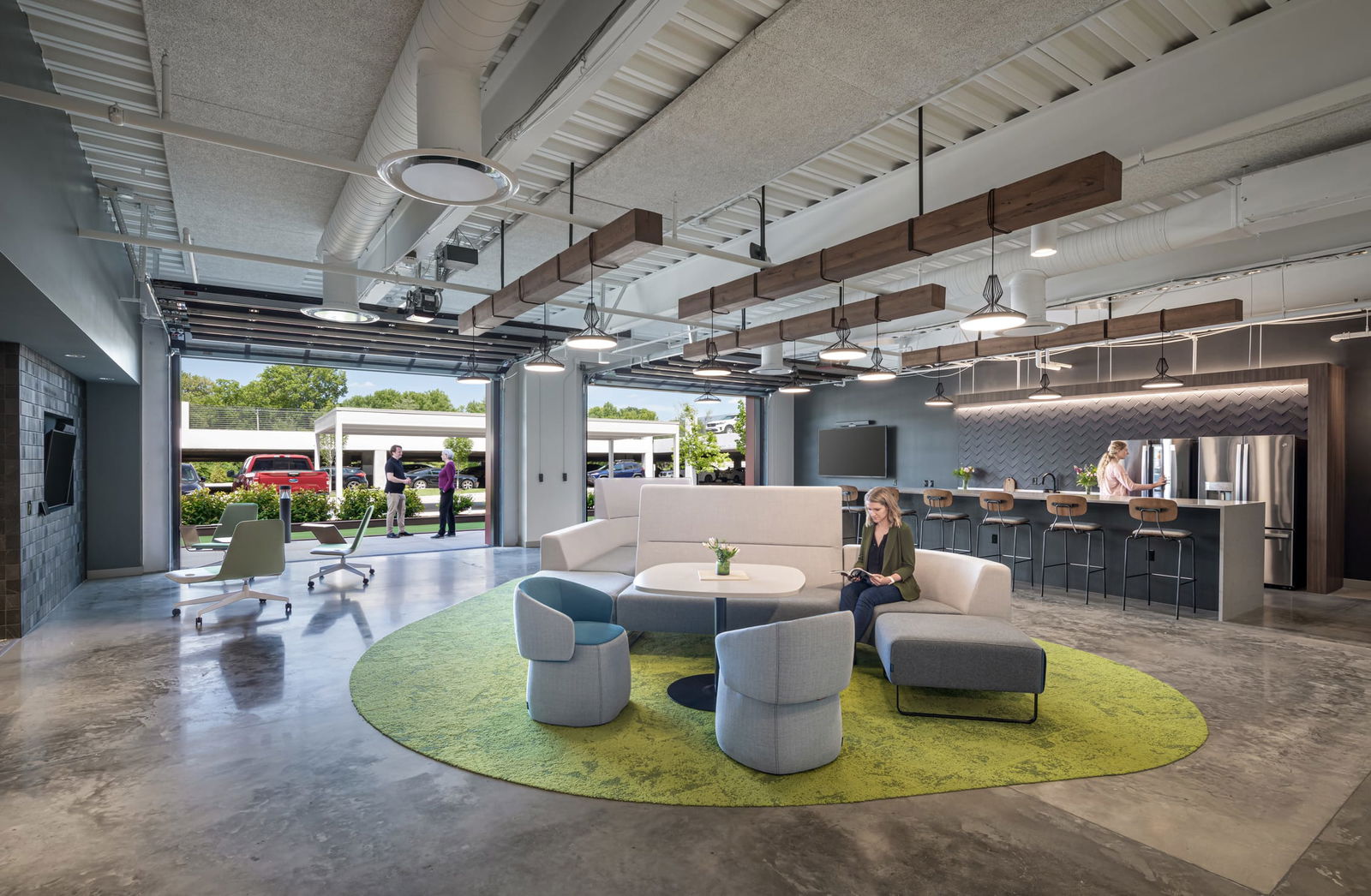 This image displays an innovative BSA-designed multipurpose space that seamlessly blends indoor and outdoor environments. The interior features a comfortable lounge area with modern curved seating in light gray upholstery arranged on a vibrant circular green area rug, where a woman sits working. Behind this lounge space is a stylish kitchen area with a dark accent wall, stainless steel appliances, and bar seating. The ceiling showcases exposed structural elements with decorative wood beams and pendant lighting. Large garage-style glass doors open completely to an outdoor space where two people are conversing, creating a flexible workspace that embraces natural light and fresh air. The polished concrete floors, modern furnishings, and thoughtful spatial arrangement demonstrate BSA's expertise in creating contemporary, adaptable work environments that encourage collaboration and wellness.