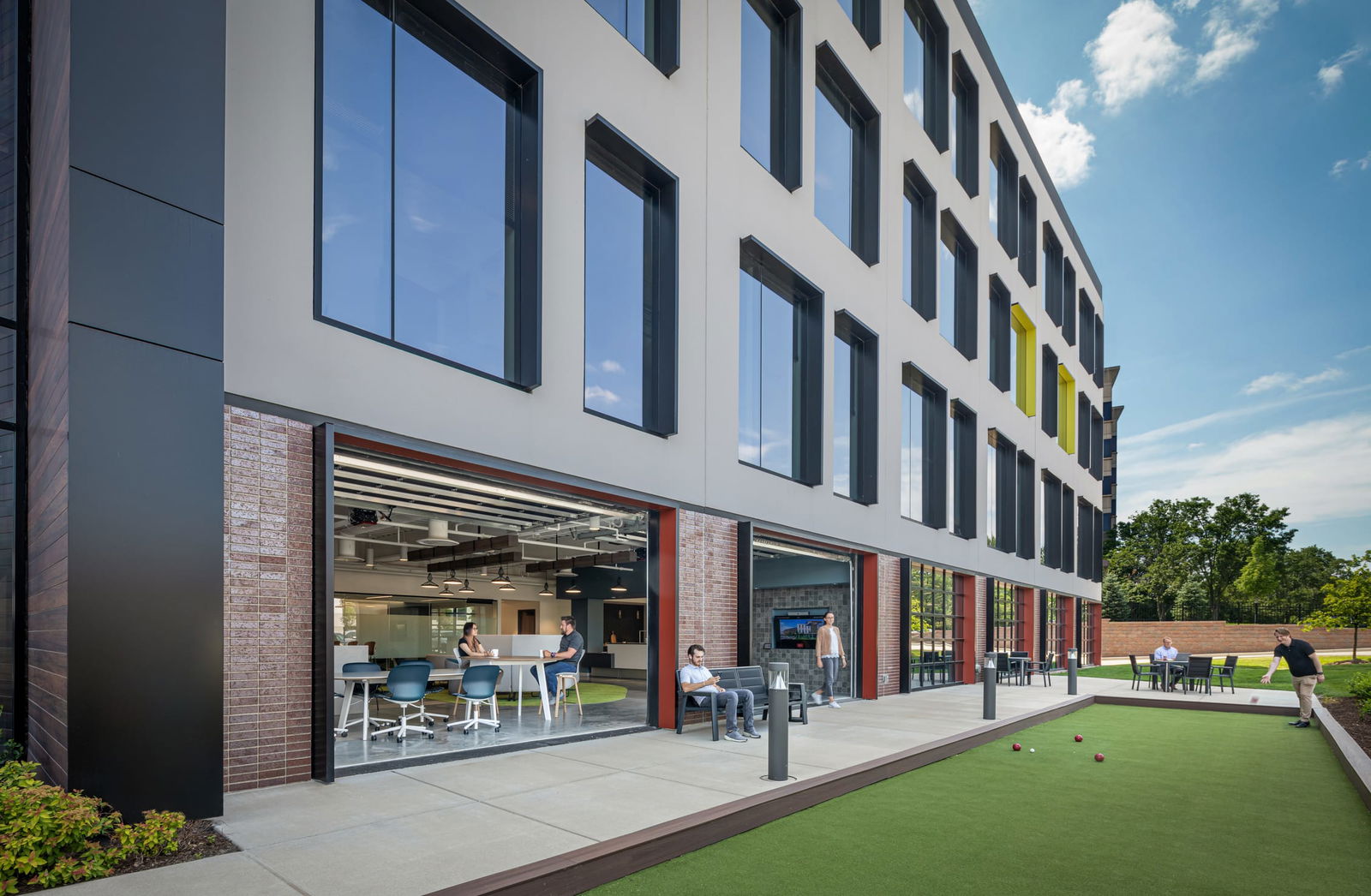 This image showcases an exterior view of BSA's office building with innovative indoor-outdoor workspace integration. The modern white façade features rhythmic black-framed windows across multiple floors, with accent elements including yellow window frames on the upper level. The ground floor opens through large garage-style doors with brick surrounds and red framing, creating a seamless transition between interior collaborative spaces and an outdoor amenity area. Inside, employees can be seen at high-top tables with modern seating. The exterior features a bocce court where someone is playing, outdoor seating areas, and landscaped elements, all under a bright blue sky. This design demonstrates BSA's expertise in creating contemporary work environments that prioritize employee wellness, collaboration, and connection to outdoor spaces.