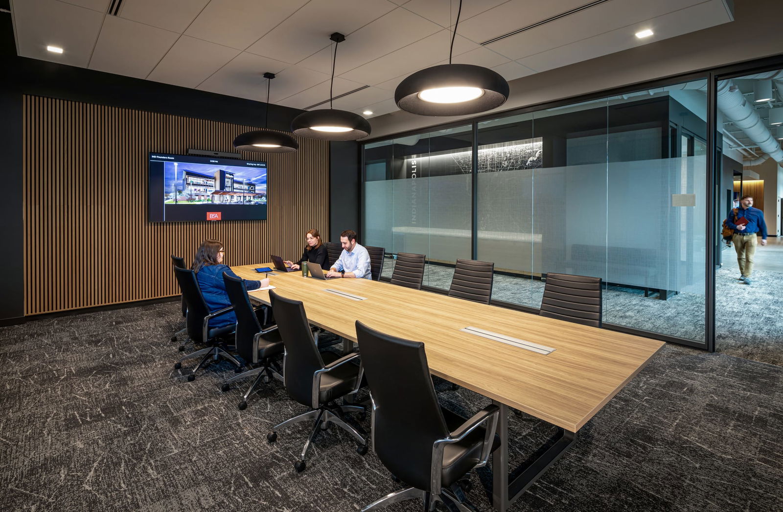 This image shows one of BSA's sophisticated conference rooms designed for collaboration and client presentations. The space features a large rectangular wooden conference table surrounded by sleek black executive chairs with metal frames. Three people are engaged in a meeting at one end of the table, working with laptops. A wall-mounted display screen showing a BSA architectural project is positioned on a feature wall with vertical wood slat paneling, creating visual warmth against the dark background. Modern disc-shaped pendant lights hang above the table, providing focused illumination. The room has glass walls with privacy frosting at eye level, maintaining openness while offering some seclusion. A person can be seen walking past in the corridor. The space demonstrates BSA's commitment to creating professional, technology-enabled meeting environments that reflect their design expertise.