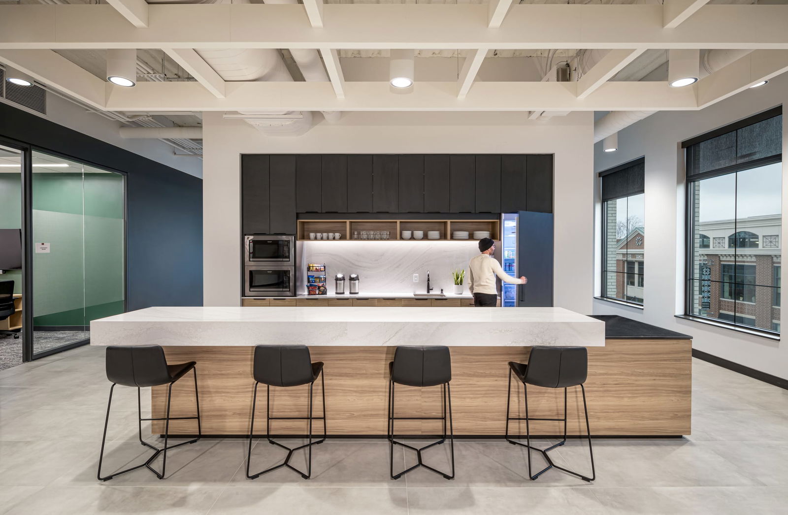 This image features BSA's modern office kitchen and break area, designed with both functionality and style. The space centers around a large island with a substantial white quartz countertop supported by warm wood cabinetry, with four contemporary black bar stools arranged along the front. Behind the island is a well-equipped kitchenette featuring dark gray upper cabinets, open shelving displaying dishware, integrated appliances, and a backlit white marble backsplash. A person can be seen accessing the refrigerator. The space benefits from natural light through large windows revealing neighboring buildings, and features an exposed ceiling with white structural elements, recessed lighting, and light-colored flooring. Adjacent glass-walled meeting spaces with accent walls are visible, highlighting BSA's thoughtful approach to creating collaborative work environments.