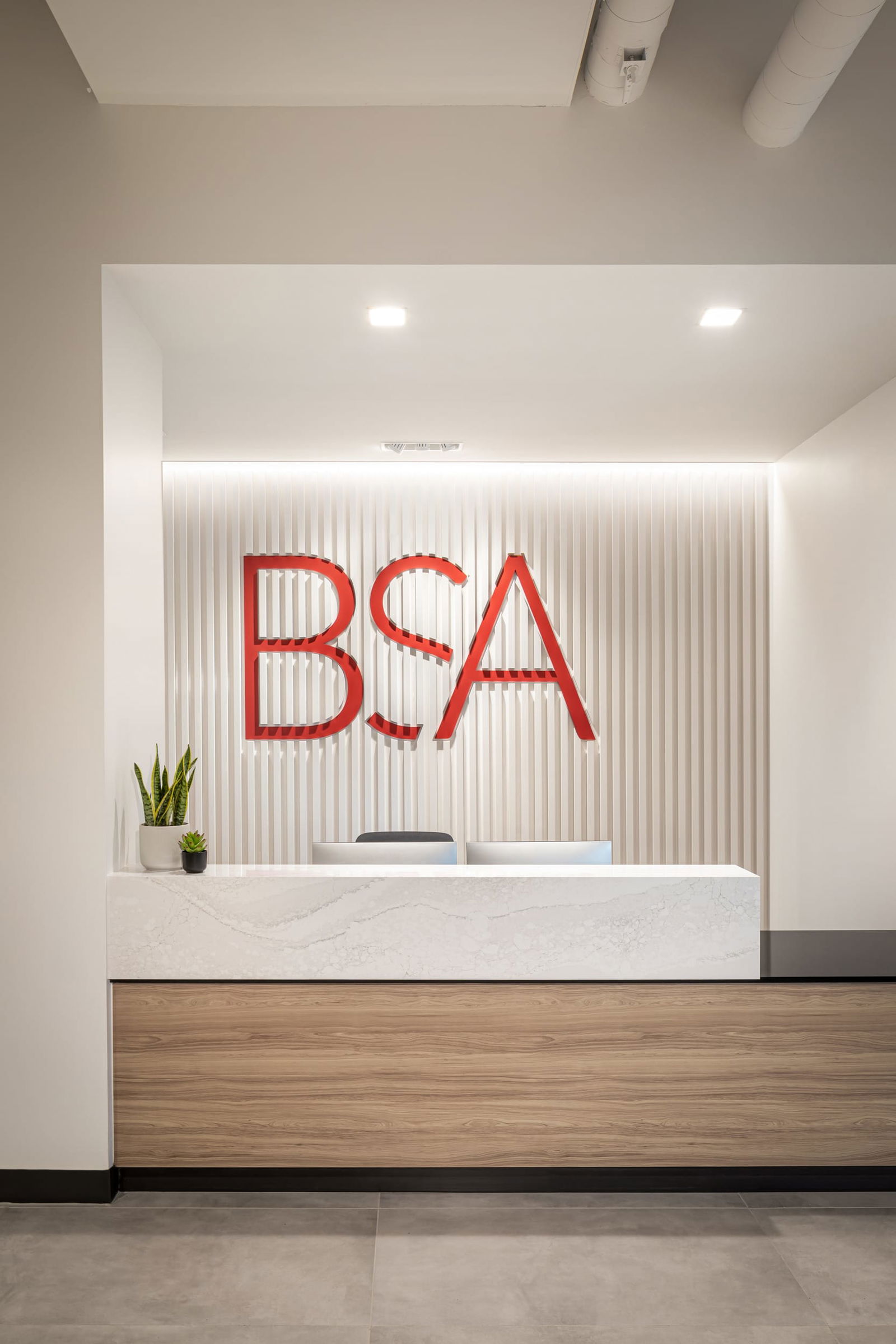 This image showcases a view of BSA's reception area, featuring a minimalist, contemporary design. The focal point is a sleek reception desk with a white marble-like top and warm wood veneer base. Behind the desk is a textured white vertical slat wall dramatically illuminated from above, with the firm's bold red