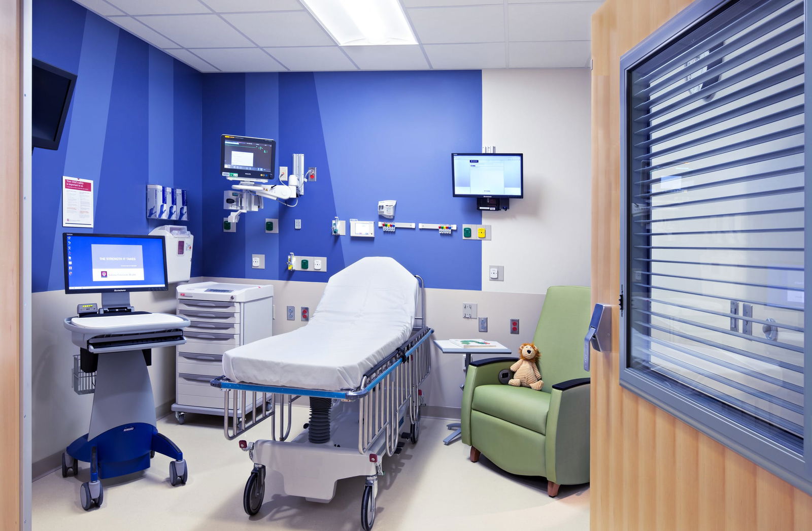 A Riley hospital room featuring a bed, a chair, a television, and a teddy bear placed on the bed. A bright blue striped mural creates a friendly and cheerful atmosphere. This image showcases BSA's expertise in designing spaces that prioritize patient care.