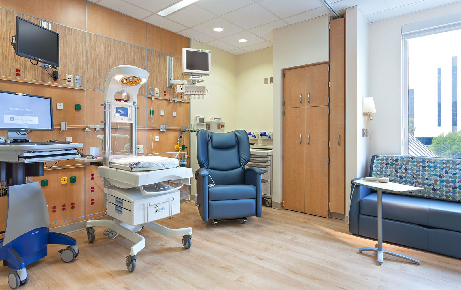 A hospital room featuring a neonatal incubator, blue reclining chair, sofa, and medical equipment, with a large window providing natural light. This image showcases BSA's expertise in designing spaces that prioritize patient care.