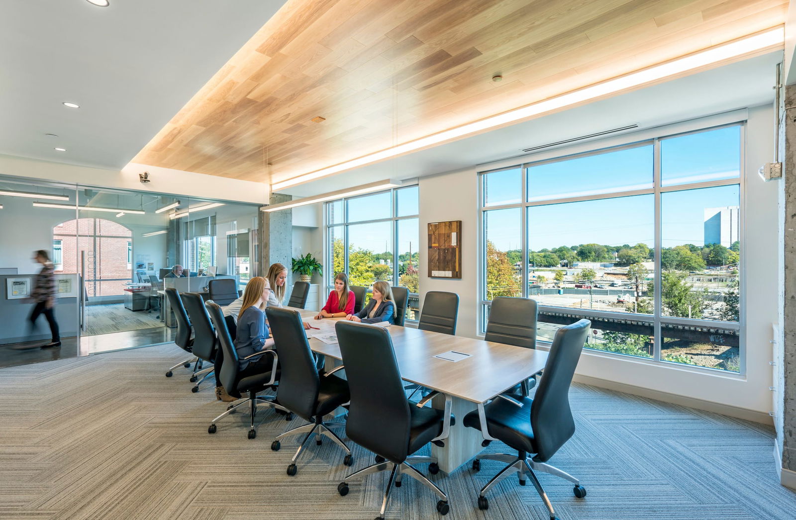 This image highlights a bright, inviting conference room in BSA’s office, reflecting the firm’s emphasis on transparency and connection. Floor-to-ceiling windows flood the space with natural light and provide panoramic views of the surrounding cityscape. The long wood-finish table and high-back ergonomic chairs create a professional yet welcoming environment for collaboration. A warm wood ceiling detail with integrated linear lighting adds texture and elegance, while adjacent glass walls maintain openness and visual continuity with the rest of the office. The design supports both focused discussion and a sense of place rooted in daylight and material richness.