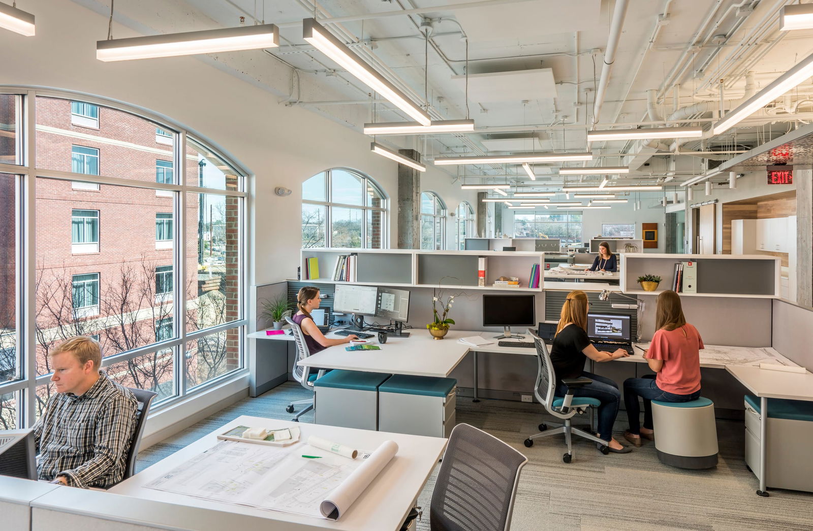 This image showcases a modern, open-concept office designed by BSA, an architecture, engineering, and interior design firm. The workspace is filled with natural light from large arched windows, creating a bright and welcoming atmosphere. The interior features clean lines, neutral tones, and pops of color through furniture and accessories. Flexible workstations, ergonomic chairs, and collaborative zones support productivity and interaction among team members. Exposed ceilings with visible ductwork add an industrial touch, while integrated greenery softens the space. The layout reflects BSA’s thoughtful approach to design, prioritizing wellness, transparency, and creativity.