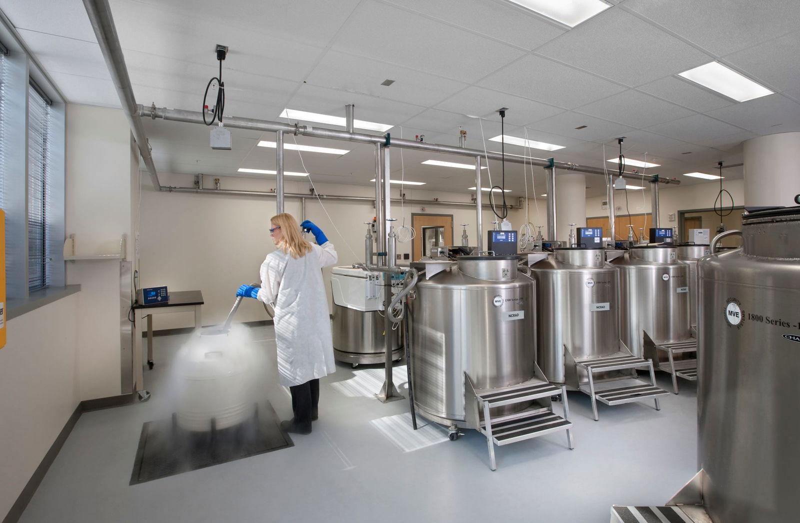 The image shows a laboratory or clean room facility with several large stainless steel tanks that appear to be bioreactors or fermentation vessels (labeled MVE 1800 Series). A person wearing a white lab coat and blue gloves is working in the space, appearing to handle something near one of the tanks. The room has a clean, clinical environment with white walls, overhead lighting, and a light-colored floor. Various utility connections hang from ceiling-mounted rails, and there are small control panels or monitors visible near some of the equipment. The space looks designed for bioprocessing, pharmaceutical manufacturing, or similar scientific applications requiring sterile conditions.
