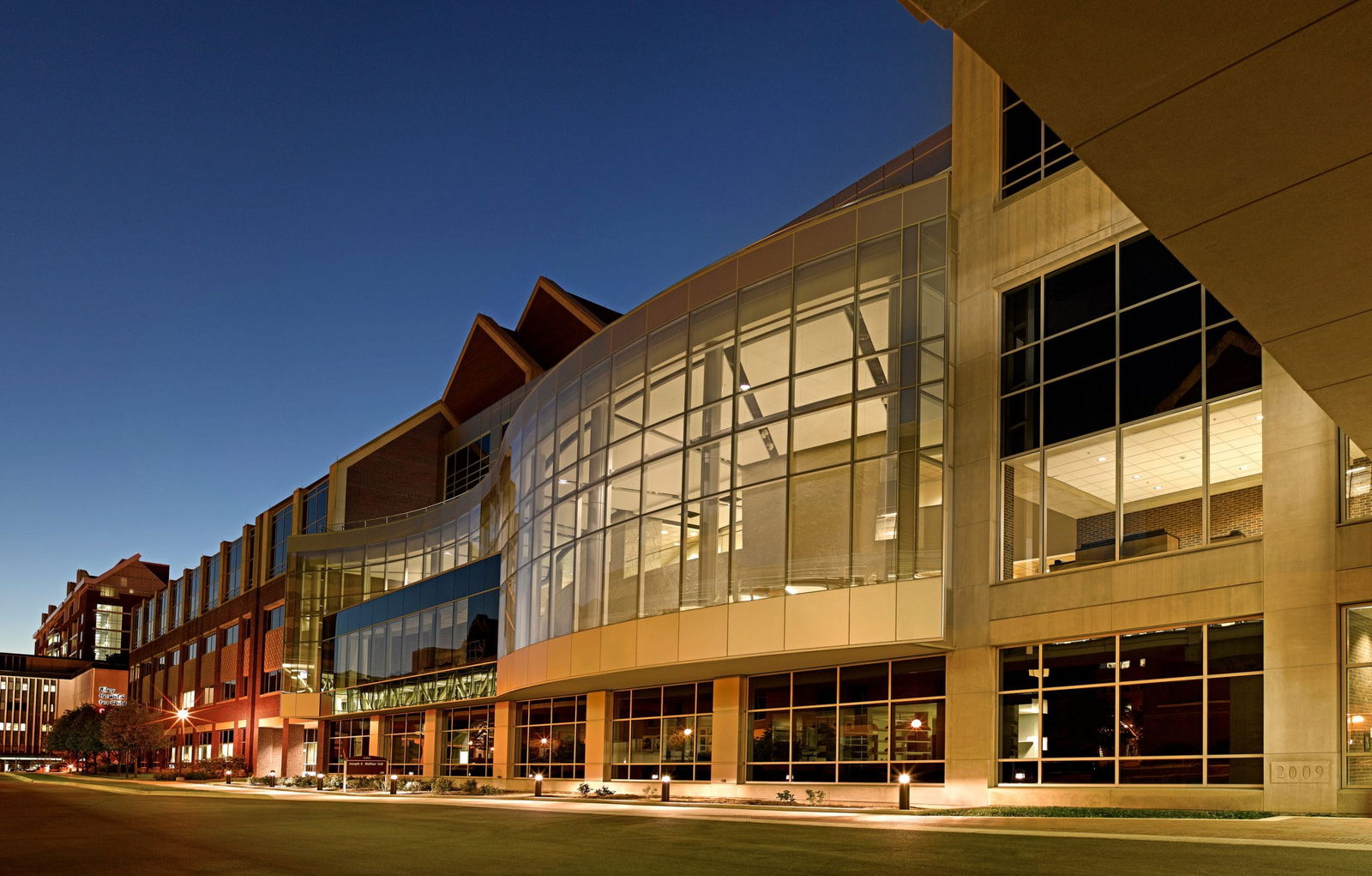 The image shows a modern institutional building photographed at twilight or early evening. The architecture features a prominent curved glass façade that creates a dramatic focal point, illuminated from within so the interior structure is partially visible. The building combines contemporary design elements with some traditional materials like brick visible on portions of the structure. The complex appears to be part of a larger campus, possibly a university science center, research facility, or corporate headquarters, given its substantial size and architectural prominence. The structure has multiple stories with varied rooflines, including some peaked elements visible at the top. The glass curtain walls reveal the interior lighting, giving the building a welcoming and active appearance despite the evening hour.