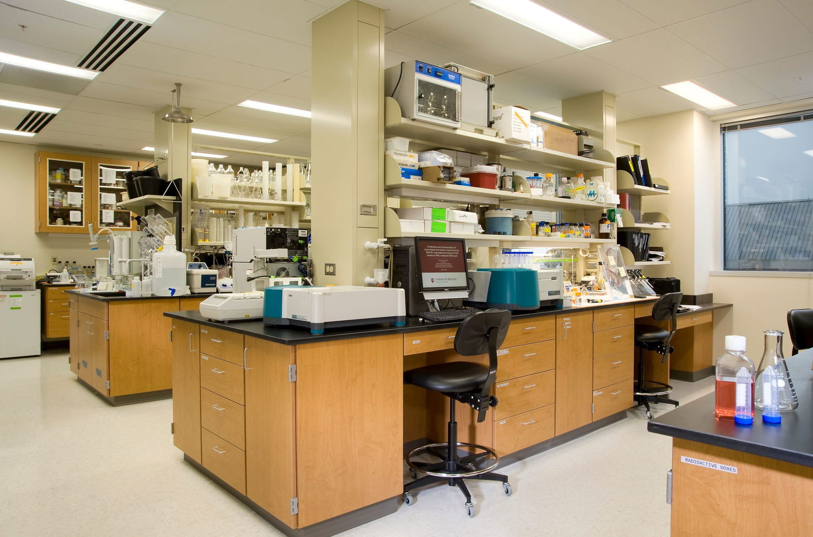 The image shows a well-equipped scientific laboratory with multiple workstations. The lab features wooden cabinetry with black countertops organized in island configurations. Several analytical instruments are positioned on the counters, including what appear to be spectrophotometers or other laboratory equipment with distinctive blue and white casings. A computer workstation is visible with a monitor and keyboard, positioned at a high desk with a black rolling lab chair. The walls have extensive open shelving holding various laboratory supplies, containers, and equipment. In the background, there's a glass-fronted storage cabinet likely containing more specialized equipment or chemicals. The space is well-lit with fluorescent ceiling lights, and a window is visible on the right side. In the foreground corner, there are several laboratory flasks containing colored liquids (one appears to be reddish and others blue). The overall appearance is of a modern, functional research or testing laboratory in an academic or industrial setting.