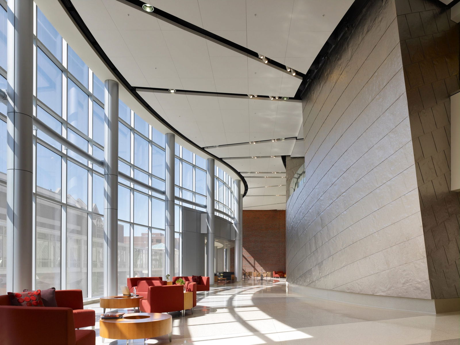 The image shows a spacious, modern architectural interior featuring a grand lobby or atrium space. The left side has a curved wall of floor-to-ceiling windows allowing abundant natural light to stream in, creating dramatic shadows across the polished floor. On the right is a large, textured stone or concrete wall with horizontal striations. The ceiling has an interesting geometric design with suspended white panels and recessed lighting. In the foreground, there are several seating areas with vibrant red armchairs and round wooden coffee tables, arranged to create conversation spaces. The overall aesthetic combines contemporary architectural elements with warm furnishings.