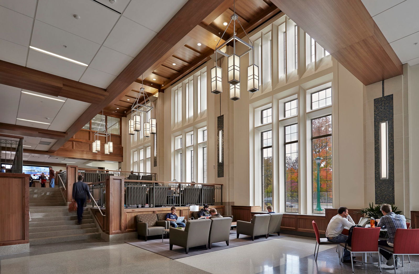 This image shows an impressive atrium or common area in the Kelley School of Business. The space features: - Dramatic two-story ceiling height with wooden architectural accents and exposed beams - Tall windows that stretch almost from floor to ceiling, allowing abundant natural light and views of autumn trees outside - Modern pendant lighting fixtures with a box/lantern design hanging from the ceiling - A split-level design with a staircase on the left leading to an upper level - A comfortable seating area with neutral-colored armchairs and sofas - Some small tables with red chairs visible in the lower right corner - People using the space for studying, socializing, and walking through The overall aesthetic combines contemporary architecture with warm wood tones, creating an inviting communal space. Several people can be seen using the area - some sitting in the lounge furniture using laptops, others at tables, and one person walking up the stairs. The architectural design emphasizes vertical space with tall windows and high ceilings, creating an open, airy atmosphere that likely serves as a gathering hub or transition space between other areas of the building.
