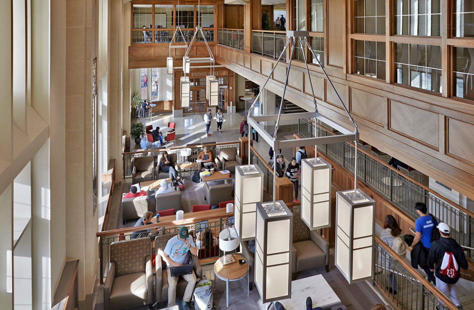 This image shows an impressive atrium or common area in the Kelley School of Business from the second floor, so that the modern pendant lighting fixtures with a box/lantern design hanging from the ceiling are eye level. The space features warm wooden architectural accents and exposed beams Below, people using the space for studying, socializing, and walking through The overall aesthetic combines contemporary architecture with warm wood tones, creating an inviting communal space.