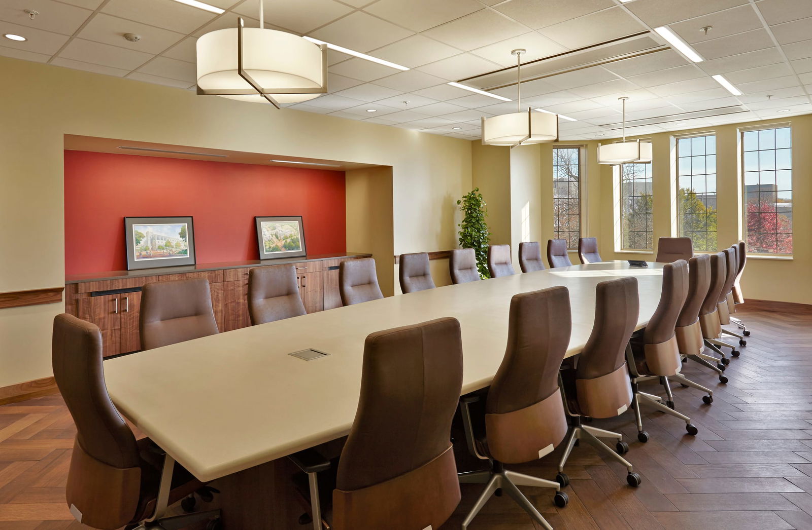 This image shows an executive style conference room with a large table and 20 leather chairs neatly arranged on all sides. The interior design incorporates neutral color palette with beige walls, wood accents, and carpeted flooring. There is one red wall that stands out amongst the rest. Large windows on one wall allow for natural light.