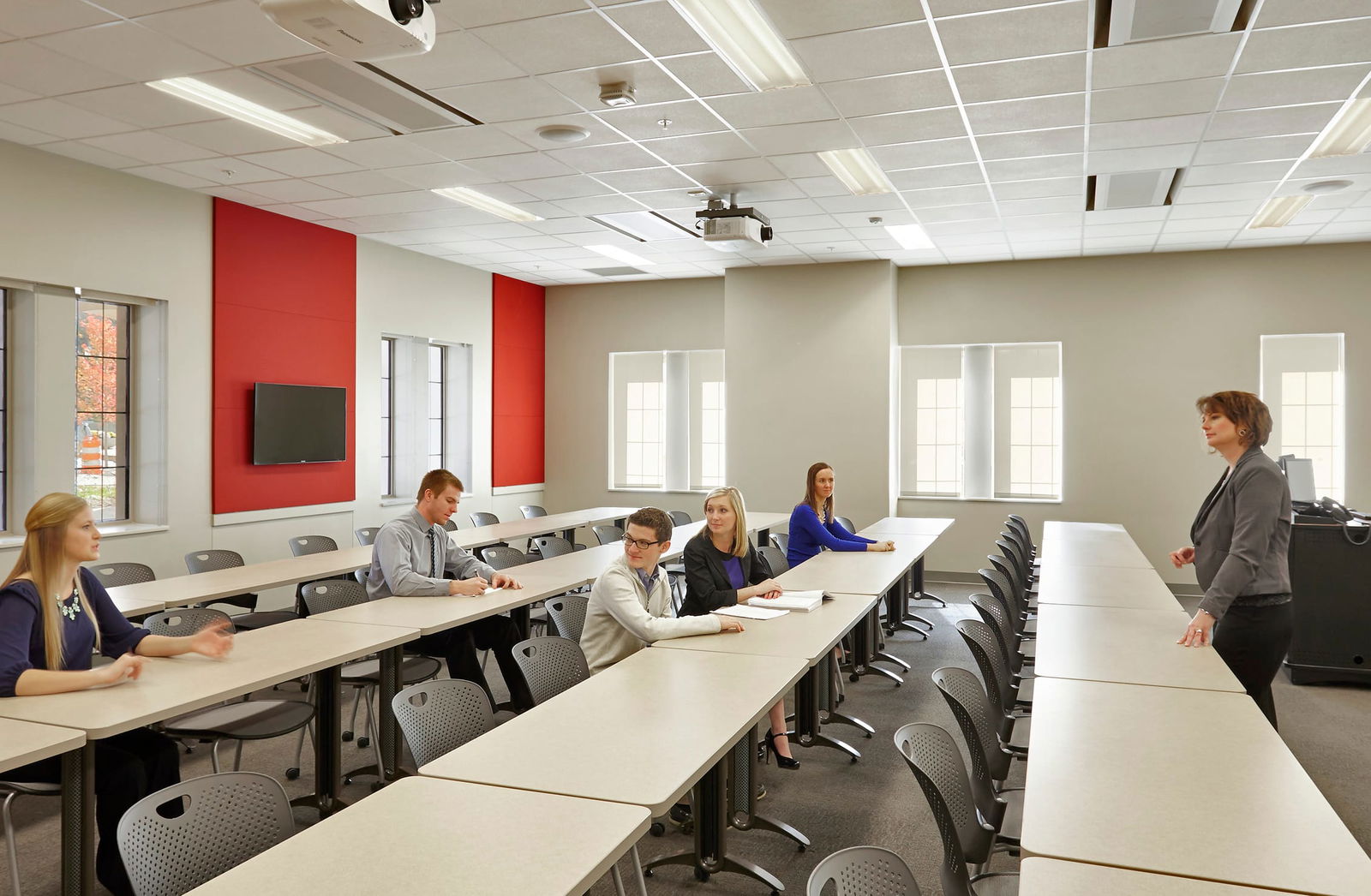 This image shows a modern classroom or lecture room. The room has several features, including long rectangular tables arranged in rows facing the front with gray plastic chairs. Red accent walls are contrasting with neutral beige/gray walls and a flat-screen TV is mounted on the red wall. Several windows provide natural light. There are several people in the room - four students or participants seated at the tables and one person (likely an instructor or presenter) standing at the front. The room has a formal academic or business training atmosphere with its organized seating arrangement.