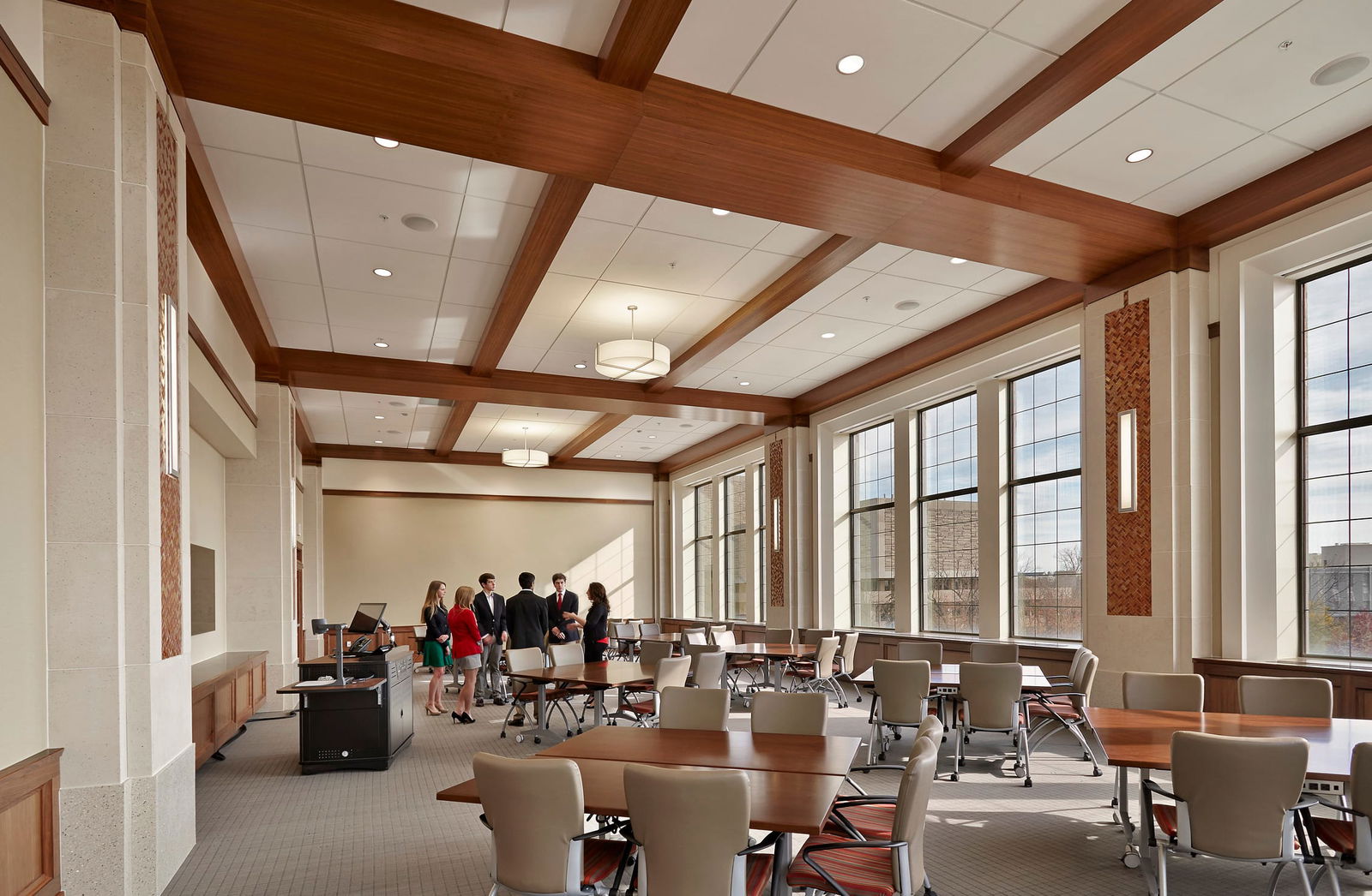 This image shows an elegant multipurpose meeting or conference room with upscale architectural elements. The space features a coffered ceiling with wooden beams contrasting against white ceiling panels and large windows along one wall allowing natural light to flood the space. Multiple tables are arranged throughout the room with cream-colored ergonomic chairs. Drum pendant lighting fixtures are suspended from the ceiling. There is a podium or presentation area at the front with what appears to be audiovisual equipment. The interior design incorporates neutral color palette with beige walls, wood accents, and carpeted flooring. In the background, a small group of professionally dressed people (approximately 5-6 individuals) are gathered in conversation. The overall aesthetic combines a traditional feel (with the wooden beams) and contemporary elements (modern furniture and lighting), creating a sophisticated space that could be used for meetings, conferences, or formal gatherings.