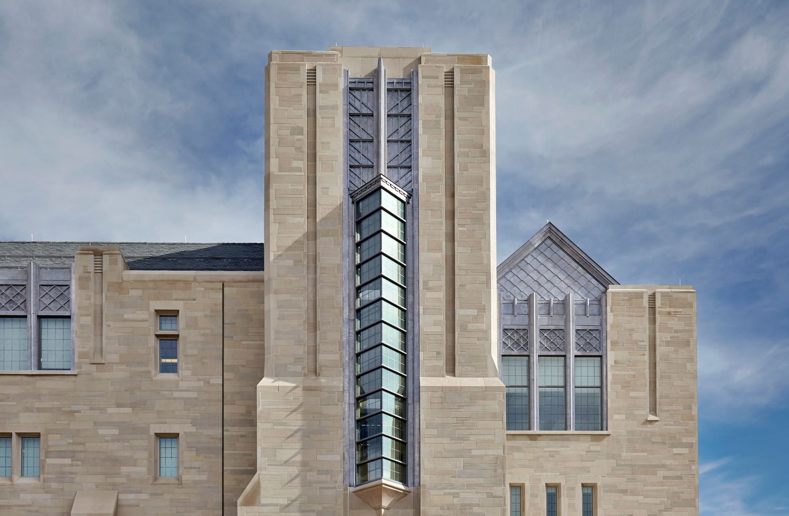 This image shows the exterior architectural detail of the Kelley School of Business. This building features Gothic Revival and contemporary design elements. The structure is built with light beige/cream limestone or sandstone blocks arranged in a uniform pattern. The most striking feature is a modern glass tower or atrium that rises vertically through the center of the building, creating a dramatic contrast between the traditional stone exterior and contemporary glass. This glass element appears to be designed to bring natural light into the interior spaces. On the right side of the building, there are large Gothic-inspired windows with diamond-pattern muntins above them. The structure features vertical stone buttresses or pilasters that add dimension and traditional collegiate Gothic character to the facade. The image is taken against a partly cloudy blue sky, which highlights the light coloration of the stone. The blend of historical architectural references with modern elements (particularly the glass tower) creates an interesting juxtaposition that respects traditional campus aesthetic while incorporating contemporary functionality.