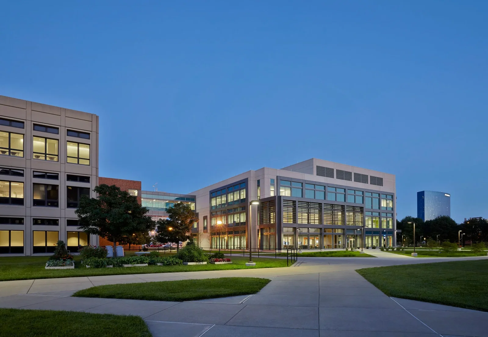 The image shows a modern institutional building (the Indiana University Science and Engineering Lab Building) photographed at dusk or early evening. The view is taken from a concrete pathway leading to the building with many other intersecting pathways to other buildings visible. The structure has a contemporary architectural design with a white exterior and extensive glass windows that are illuminated from within, creating a warm glow against the deep blue sky. To the left is another building with a similar institutional design, and between them is a landscaped area with green lawns. The scene is peaceful with no people visible, and the lighting suggests it's after business hours when the buildings are still operational but the campus is quiet.