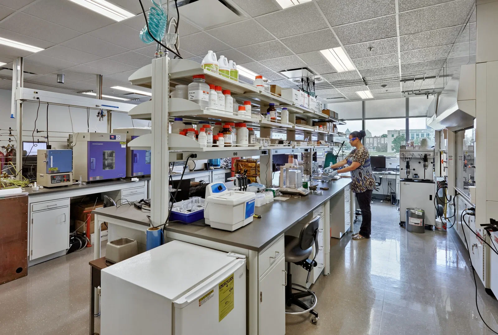 This image shows a professional research laboratory with advanced scientific equipment. The lab features long workbenches with various analytical instruments and a person working at one of the stations wearing protective eyewear. Above the central bench are several shelves stocked with numerous chemical reagents and supplies in bottles with colored caps. The space includes specialized equipment like what appears to be ovens or incubators with purple doors on the left wall, and various analytical instruments distributed throughout the workspace. There's a refrigerator or cold storage unit in the foreground, and the lab has proper utility connections hanging from the ceiling. Large windows at the far end allow natural light and show another building outside. The polished floor and organized layout suggest this is a well-maintained, modern research facility, likely in a university or pharmaceutical/biotech setting.