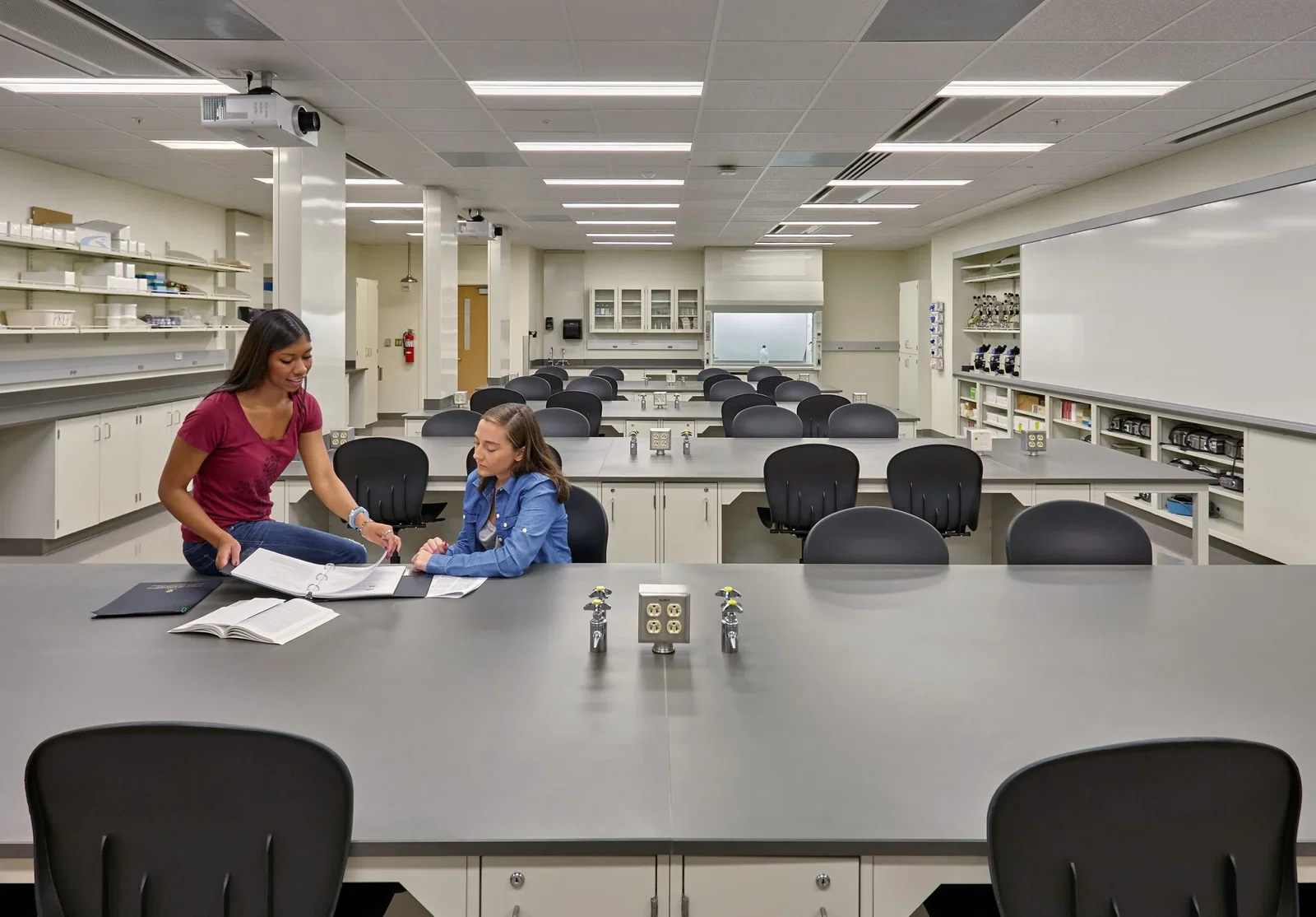 This image shows a modern science laboratory classroom. Two students are collaborating at one of the lab benches - one wearing a burgundy top and the other in a denim jacket - reviewing materials in a binder or notebook. The room features multiple lab stations with black chairs and gray countertops equipped with utility connections visible in the center of each table. Around the perimeter, there are storage cabinets, shelving units containing laboratory supplies and equipment, and what appears to be a fume hood or safety station at the back. The space is well-lit with fluorescent ceiling lights, and a projector is mounted on the ceiling. The clean, organized environment suggests this is a newly built or recently renovated teaching laboratory designed for hands-on science instruction.