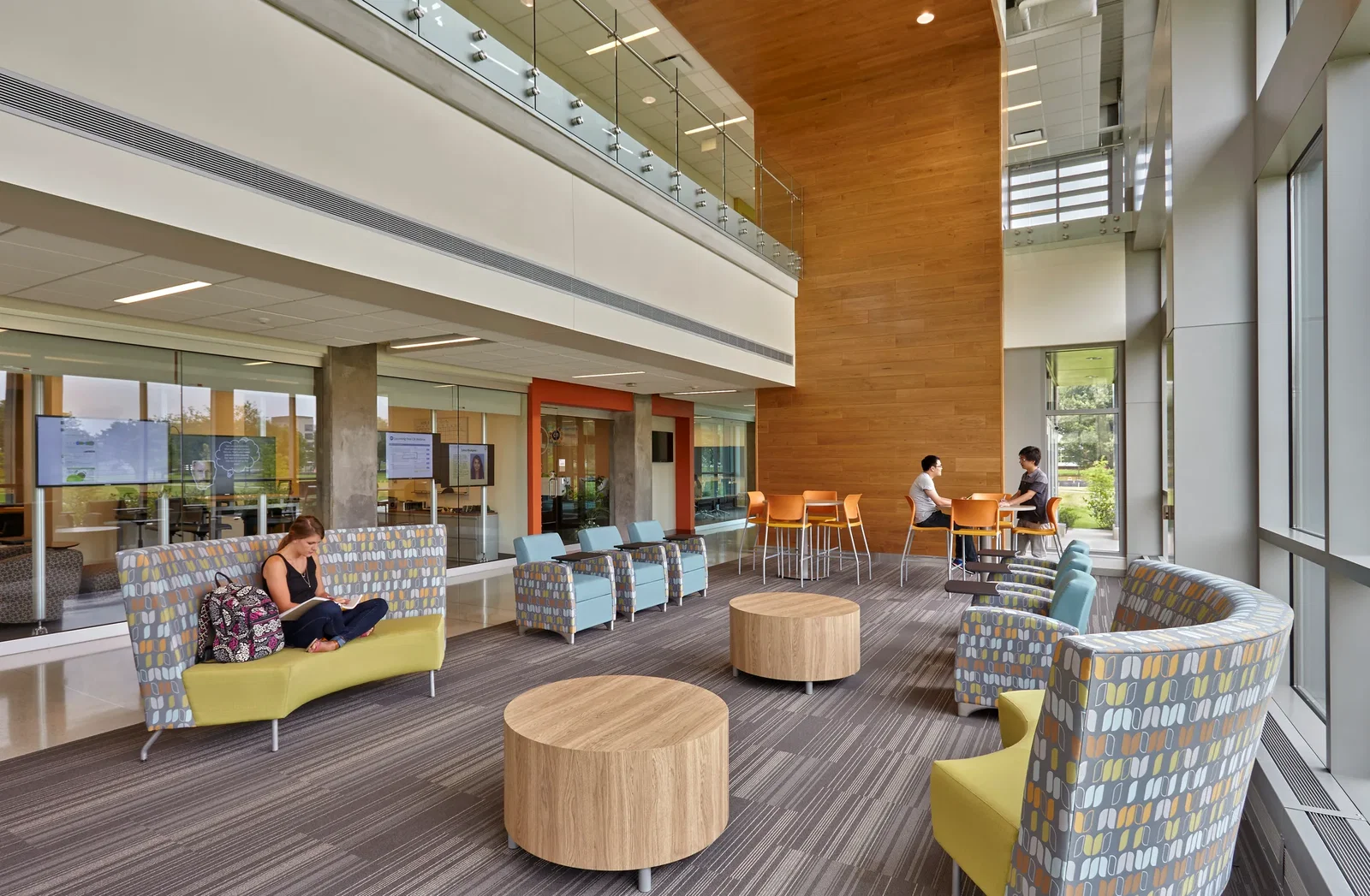 This image shows a modern, open common area in the IU Science and Engineering Laboratory Building. The space features a two-story design with a mezzanine level visible at the top, protected by glass railings. In the foreground, there's a comfortable seating area with curved sofas upholstered in a mid-century modern pattern with blue, yellow and gray geometric shapes. The sofas have bright yellow accents, and there are wooden cylindrical coffee tables positioned in the center of the seating arrangement. The floor has a striped gray carpet pattern. On the left side, a person with darker hair is sitting on one of the sofas, appearing to read or work. To the right, in the background near a prominent wooden wall feature, two people are sitting at a high table having a conversation. The space is bright and airy with large windows on the right side allowing natural light and views of greenery outside. There are also glass-walled rooms visible in the background, likely meeting rooms or study spaces. The overall design combines white walls, warm wood elements, and pops of color in the furniture to create an inviting, contemporary atmosphere for collaboration or focus.