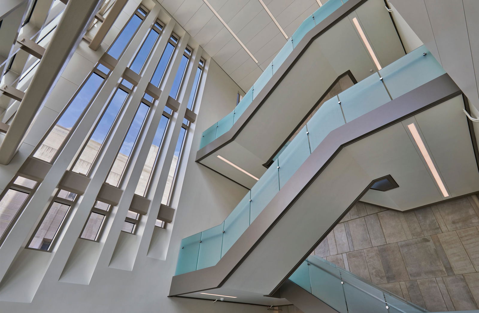 This image shows a striking architectural interior featuring a modern atrium space. The photograph is taken looking upward, capturing multiple floors with angular, geometric design elements. On the left side, there's a dramatic wall of tall, vertical windows with white architectural fins or supports that create a rhythmic pattern. The windows allow natural blue sky to be visible, bringing daylight deep into the building. On the right side of the image, there are several floors of balconies or walkways with minimalist, contemporary design. These balconies feature frosted glass railings with a light blue-green tint, supported by metal fixtures. The walkways appear to follow a geometric, angular pattern rather than traditional curved forms. The ceiling is white with integrated lighting, and the overall color palette of the space is dominated by whites, light grays, and the subtle blue tint of the glass. Some concrete or stone textured surfaces are visible in portions of the walls. This photo shows architectural design principles with emphasis on light, openness, and geometric forms.