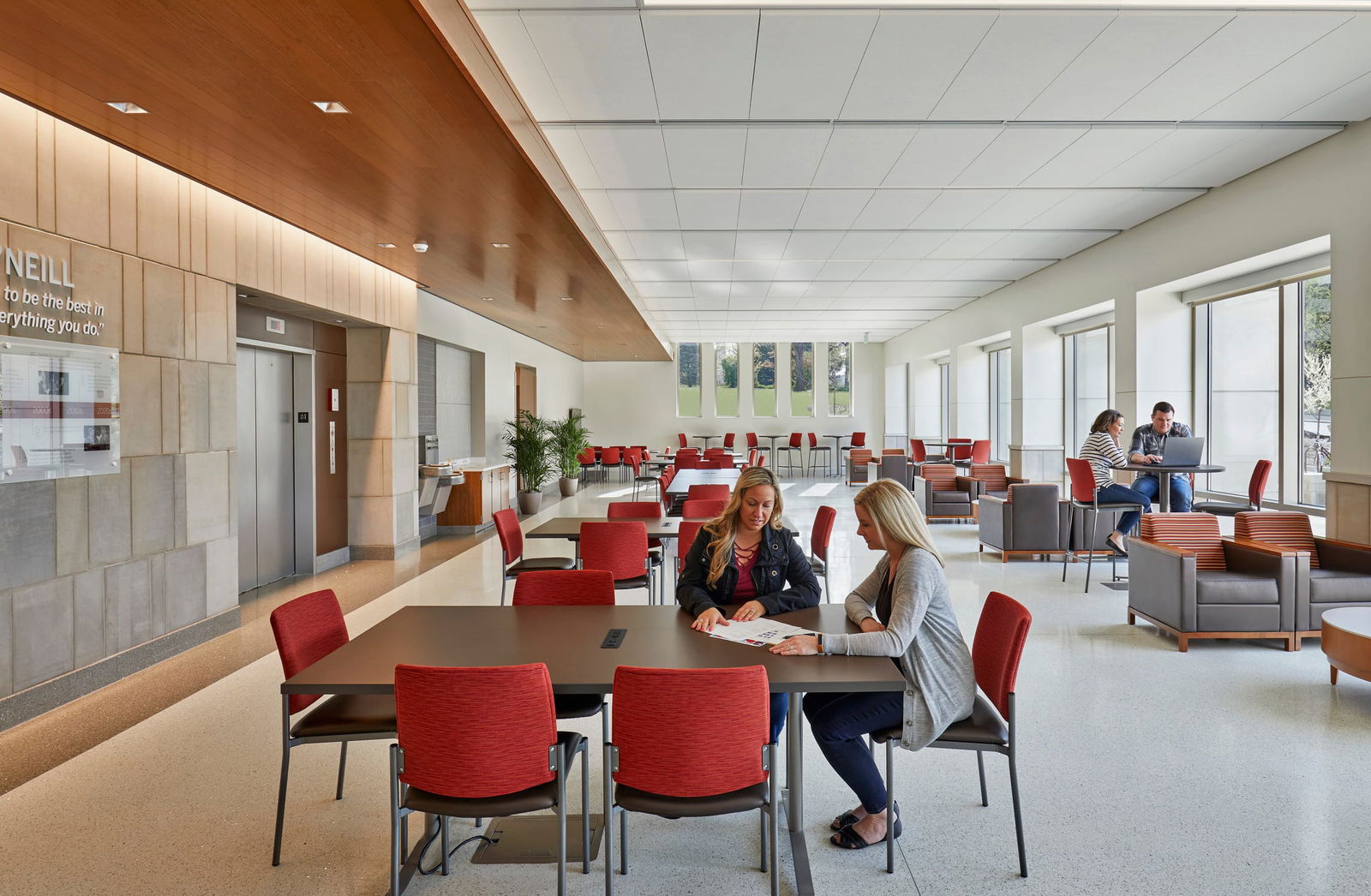 The image shows a modern, open-concept student workspace with a sleek, contemporary design. In the foreground, there's a large wooden communal table with bright red chairs. Two women are working collaboratively at this table over a piece of paper. The space extends back to reveal multiple similar seating arrangements, where a few people can be seen working. The area features floor-to-ceiling windows along the right side, bringing in natural light and providing views of trees outside. The interior combines warm wood elements with clean, neutral tones including white ceilings, glass partitions, and gray flooring with a subtle pattern. The space appears designed for collaboration, with a mix of communal tables and some lounge seating with armchairs visible toward the back.