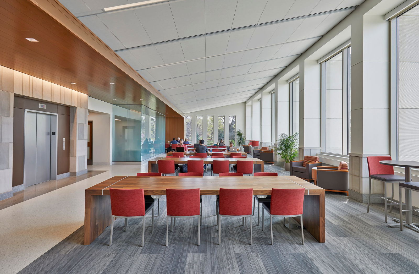 The image shows a modern, open-concept student workspace with a sleek, contemporary design. In the foreground, there's a large wooden communal table with bright red chairs. The space extends back to reveal multiple similar seating arrangements, where a few people can be seen working. The area features floor-to-ceiling windows along the right side, bringing in natural light and providing views of trees outside. The interior combines warm wood elements with clean, neutral tones including white ceilings, glass partitions, and gray flooring with a subtle pattern. To the left, there's an elevator area with wood paneling and glass walls. The space appears designed for collaboration, with a mix of communal tables and some lounge seating with armchairs visible toward the back. Green plants add a touch of nature to the environment, contributing to a fresh, welcoming atmosphere.