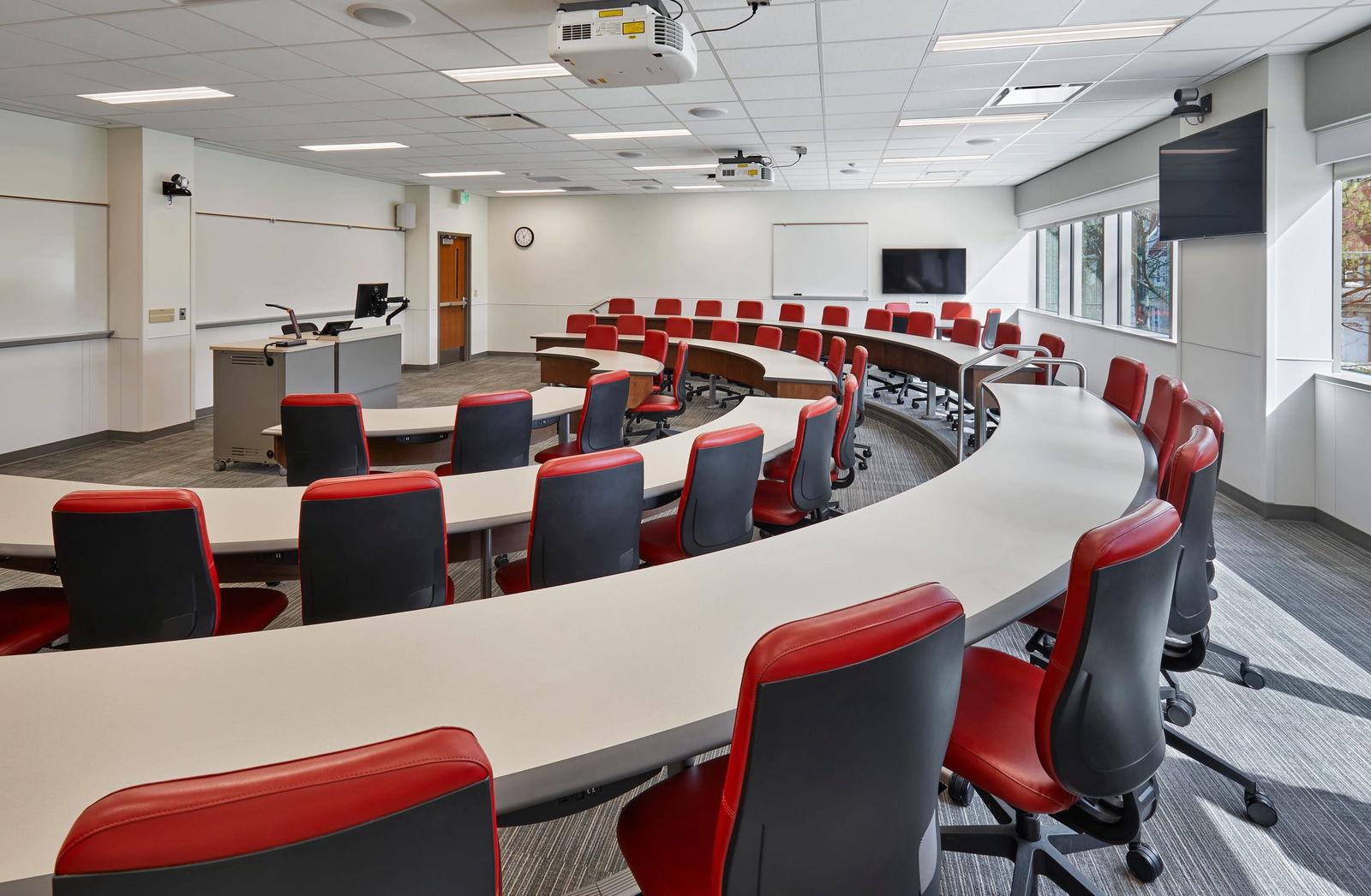 This image shows a modern educational classroom designed in a tiered, semi-circular layout. The room features several curved rows of white tables arranged in an amphitheater style, with red and black office chairs at each workspace. This design allows students to face toward the front of the room where there's an instructor's desk or podium equipped with a computer. The classroom is outfitted with educational technology including ceiling-mounted projectors, a wall-mounted TV display, and a whiteboard. Windows along one side provide natural light, and there are window blinds that can be adjusted for presentations. The space has a professional, contemporary feel with neutral walls, carpeted flooring with a gray pattern, and a drop ceiling with integrated lighting. A wall clock and door to the hallway are visible in the background. This type of classroom is designed to facilitate discussion and ensure good visibility for all participants.