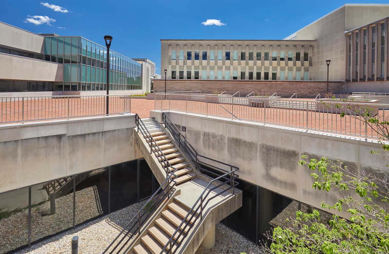 This image shows a modern institutional campus with brutalist architectural elements. The composition features several concrete structures arranged around a multi-level exterior space. In the foreground, there's a prominent concrete staircase with metal railings that connects different levels of what appears to be a sunken courtyard or light well area. The lower level contains gravel or small stones. The upper level consists of a brick-paved plaza or courtyard with metal railings along its perimeter. Multiple academic buildings surround this courtyard - the most prominent being a rectangular structure in the center background with a distinctive façade featuring vertical concrete elements alternating with windows. To the left, there's a more contemporary section with extensive glass curtain walls. The overall aesthetic combines utilitarian concrete architecture with more modern design elements, typical of university or institutional campuses built or renovated across different eras.
