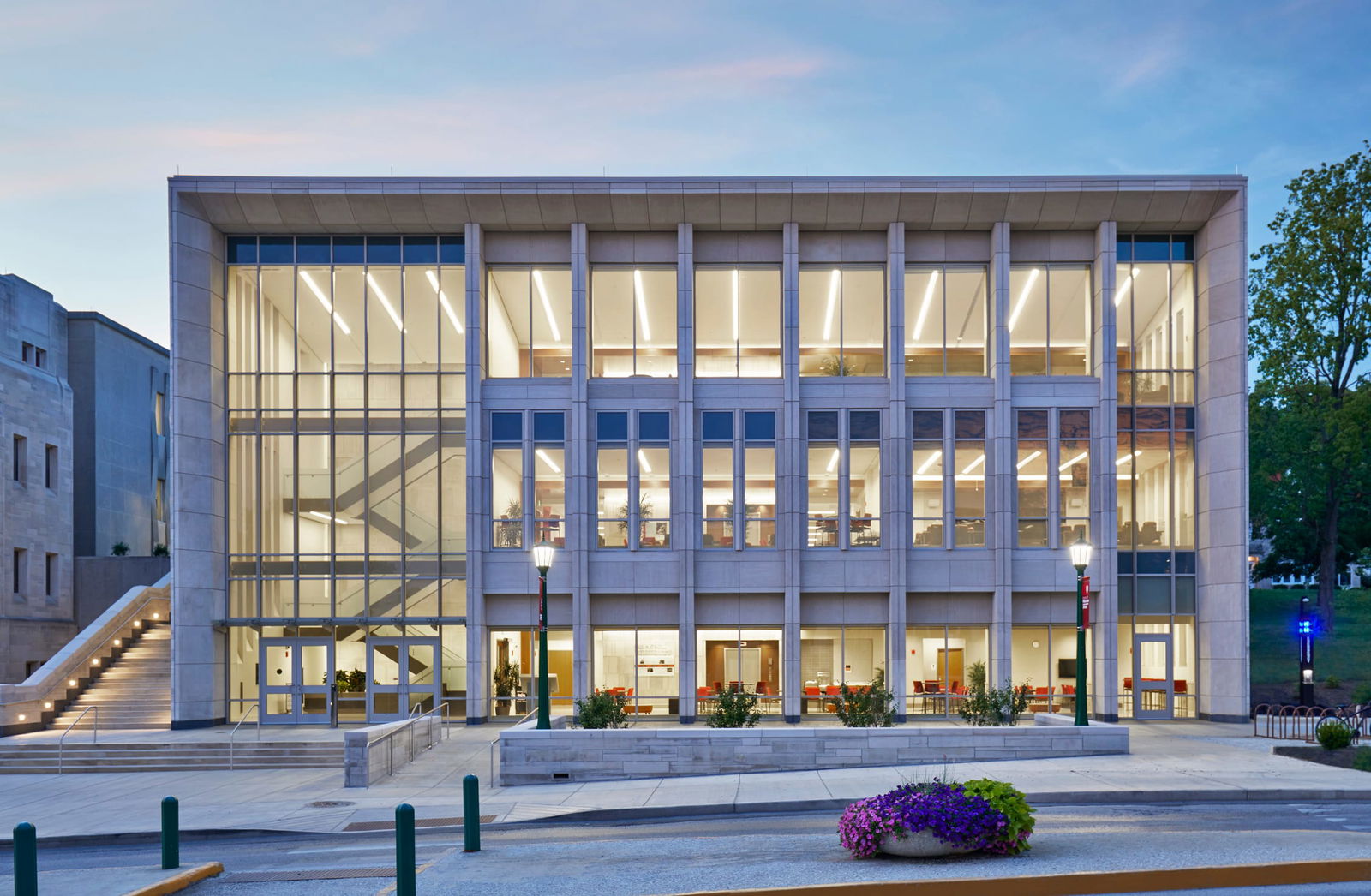 This image shows a modern academic building on IU Bloomington's campus photographed at dusk or early evening. The structure features a contemporary design with an extensive glass façade that reveals three illuminated floors inside. The building is constructed with limestone with vertical architectural elements framing the large glass windows. Through the glass, interior spaces with furniture are visible, suggesting study areas or collaboration spaces. A prominent staircase can be seen through the glass on the left side. The building has an exterior staircase with handrails on the far left, leading to an upper entrance. Lamp posts with architectural lighting fixtures illuminate the entrance.