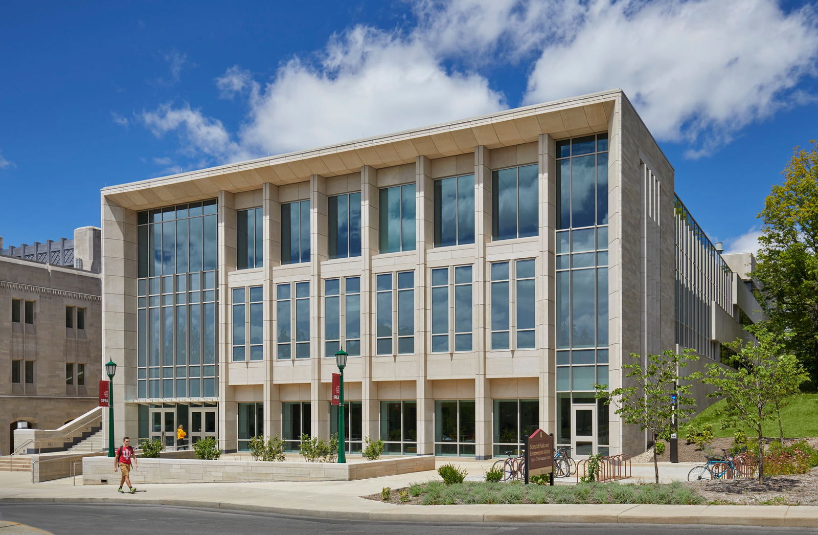 This image shows a modern academic building on IU Bloomington's campus photographed against a beautiful blue midday sky. The structure features a contemporary design with an extensive glass façade that reveals three illuminated floors inside. The building is constructed with limestone with vertical architectural elements framing the large glass windows. The building has an exterior staircase with handrails on the far left, leading to an upper entrance.