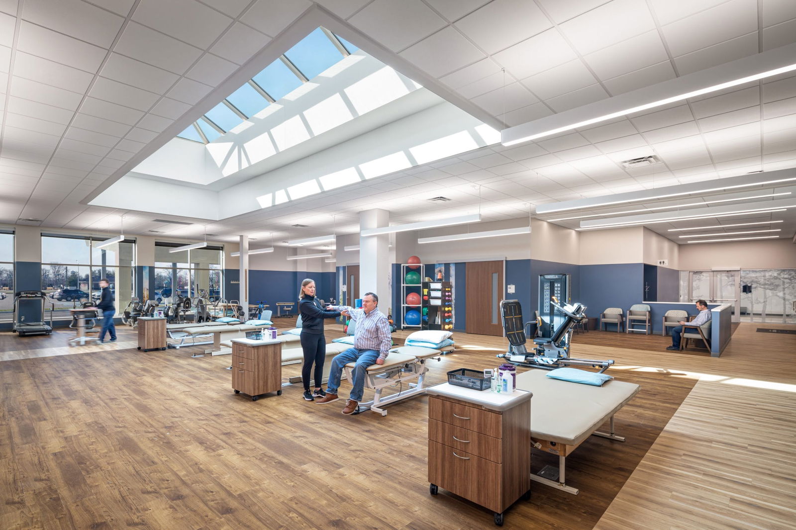 Physical therapy or rehabilitation room within the converted mall healthcare facility. A large geometric skylight floods the spacious room with natural light. Examination tables, medical equipment, and wooden cabinets are arranged throughout the space. A healthcare professional is talking with a patient seated on an examination table. The room has wood-tone flooring, white walls, and blue accent panels. Medical exercise equipment and other treatment stations are visible in the background.