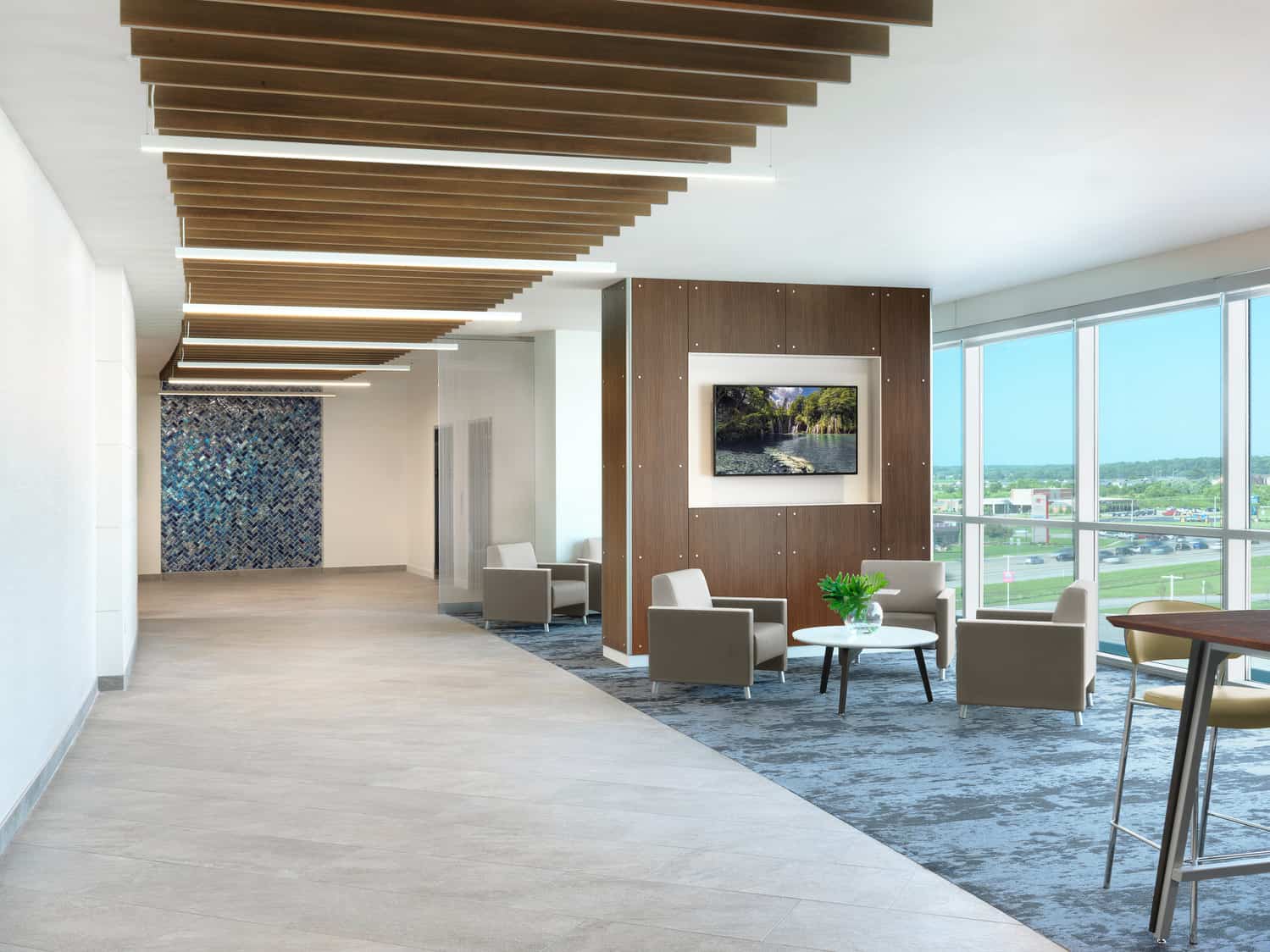 A long hallway within the Deaconess Hospital with a patient family waiting area on the right side. Sets of brown and green seating surrounding tables are separated by entertainment systems. Floor-to-ceiling windows allow natural light to flood the space.
