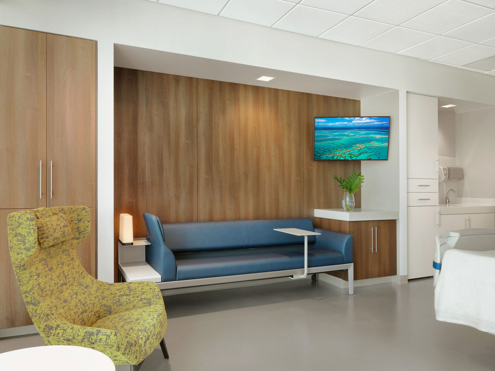 Interior of a modern private patient room for overnight hospital stays, including a blue couch and green chair in front of a wood paneled wall. This would be the view of the patient in the bed.