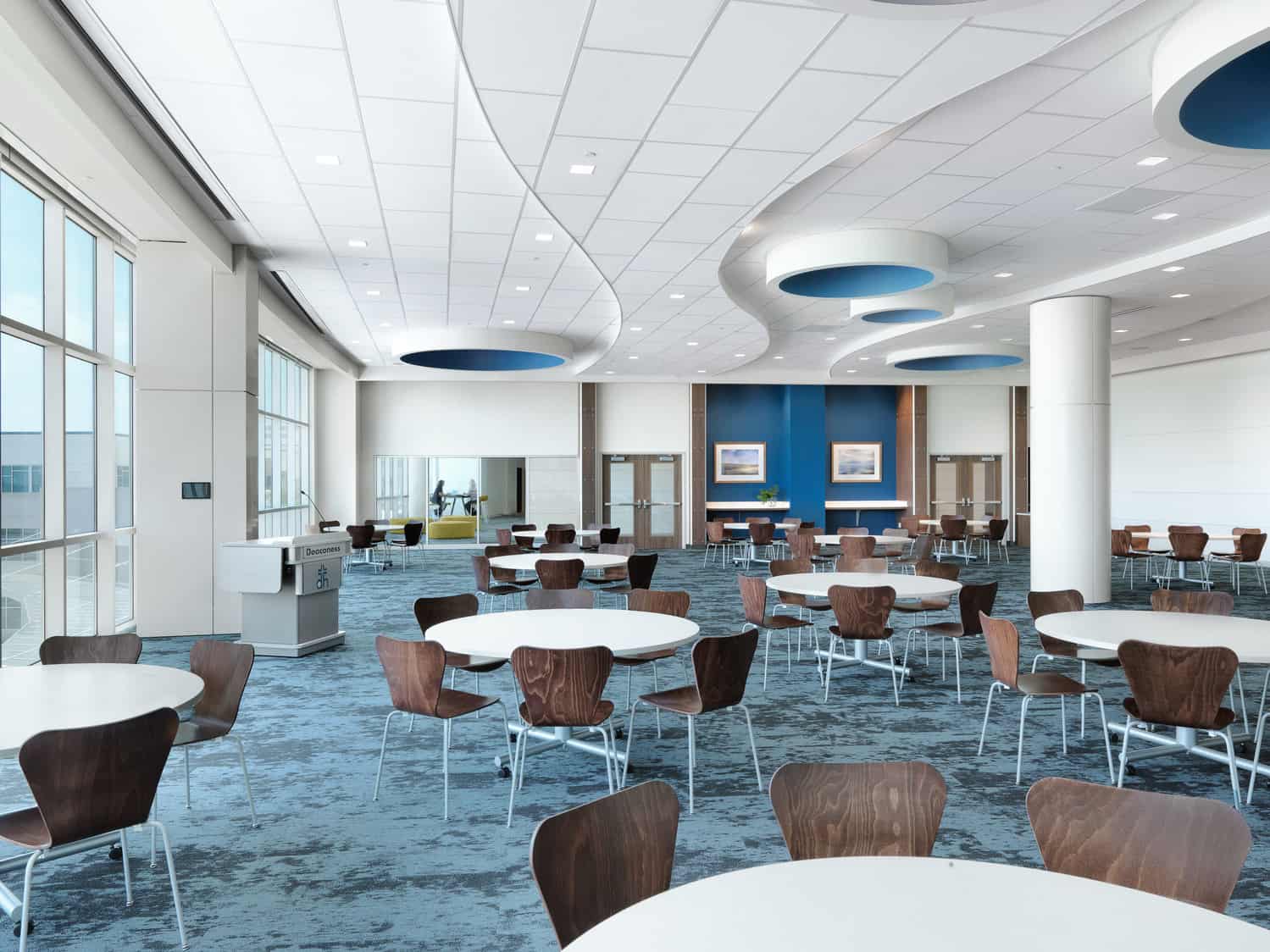 A conference-like room with blue carpet and white tables with modern, dark wooden chairs are arranged around a podium, The ceiling is paneled and features waves at different elevations. This room shows BSA's expertise in interior design.