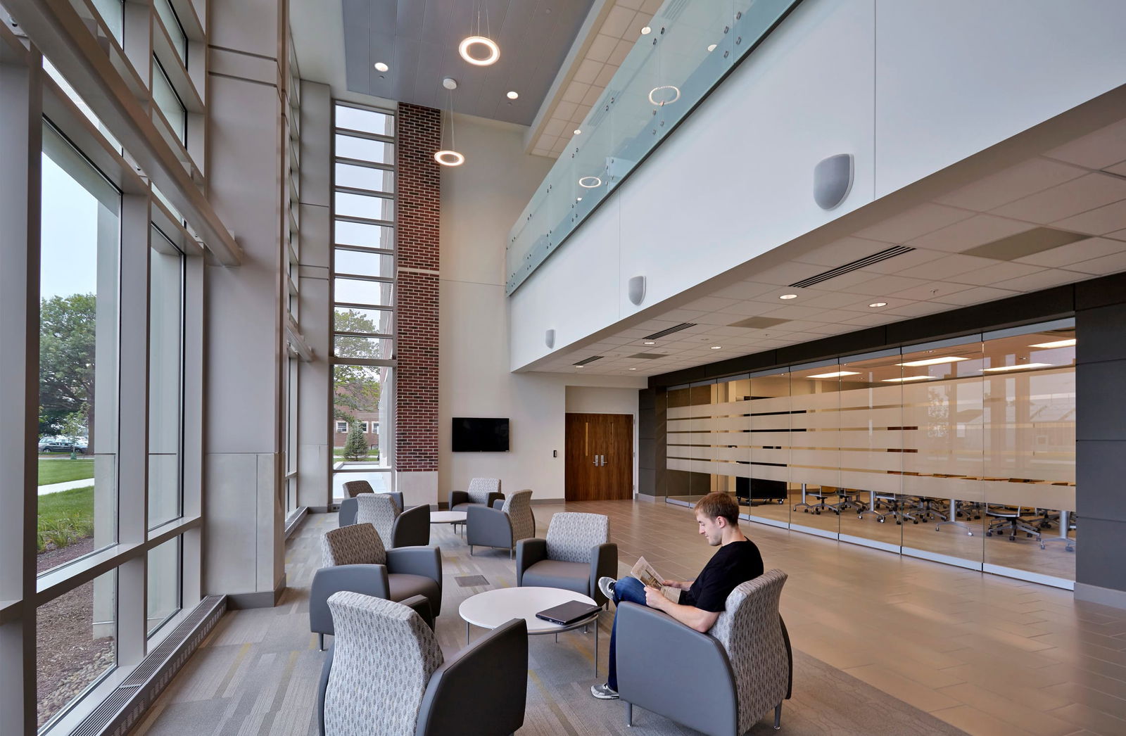 A spacious, modern lounge area within the Purdue Institute for Drug Discovery with high ceilings and floor-to-ceiling windows that allow ample natural light. The space is furnished with gray upholstered armchairs arranged around small circular tables. A man in a black T-shirt and jeans sits in one of the chairs, reading a book with a laptop on the table beside him. The interior features a mix of brick, glass, and sleek white walls. A glass-walled conference room with horizontal frosted stripes is visible in the background, along with a mezzanine level with a glass railing above. Pendant lights hang from the ceiling, enhancing the contemporary aesthetic.