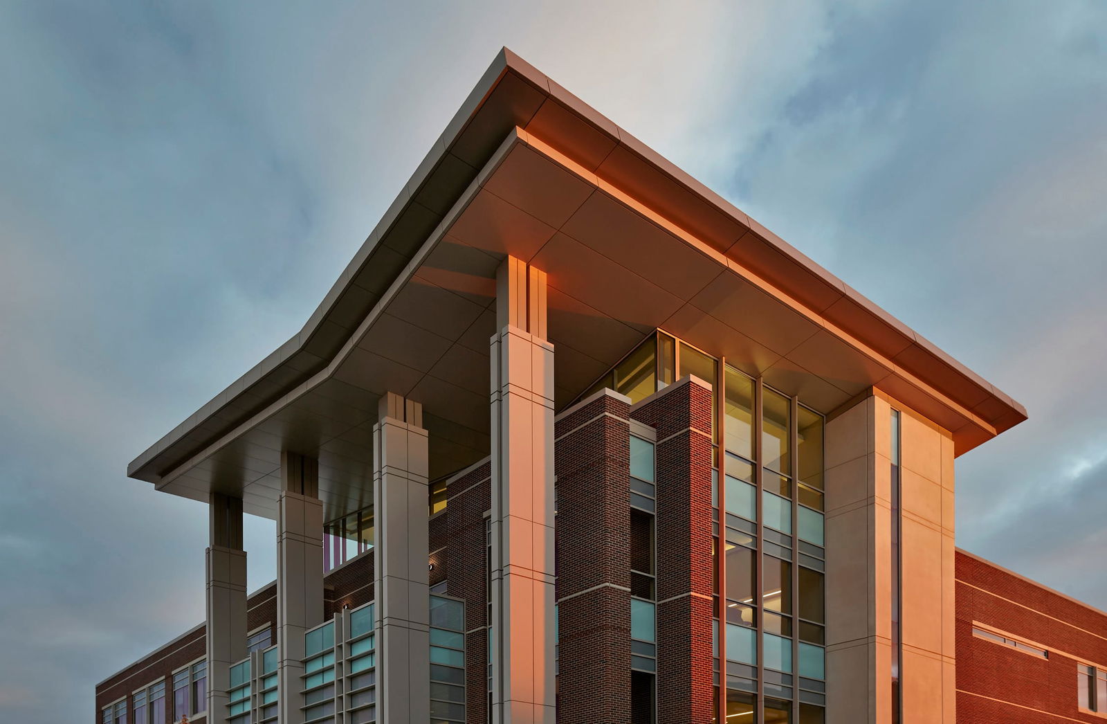 A modern academic building captured at sunset, with warm sunlight casting a golden glow on its architectural features. The structure combines brick, glass, and metal elements, with tall, rectangular columns supporting an extended roof. Large windows reflect the sky, while sleek metal accents and horizontal louvers add to the contemporary design. The background features a partly cloudy sky, adding depth and contrast to the image.