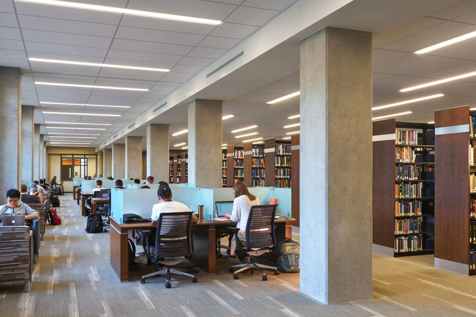 A modern academic library in the Purdue University Active Learning center with rows of study carrels occupied by students working on laptops and reading. The space features a combination of individual study desks with frosted glass dividers and open seating areas. Tall concrete columns support the ceiling, which has a grid of recessed lighting. Wooden bookshelves filled with books line the right side of the image, while students are scattered throughout, focused on their work. Some students have backpacks and personal items at their desks. The atmosphere is quiet and studious, with a mix of natural and artificial lighting.