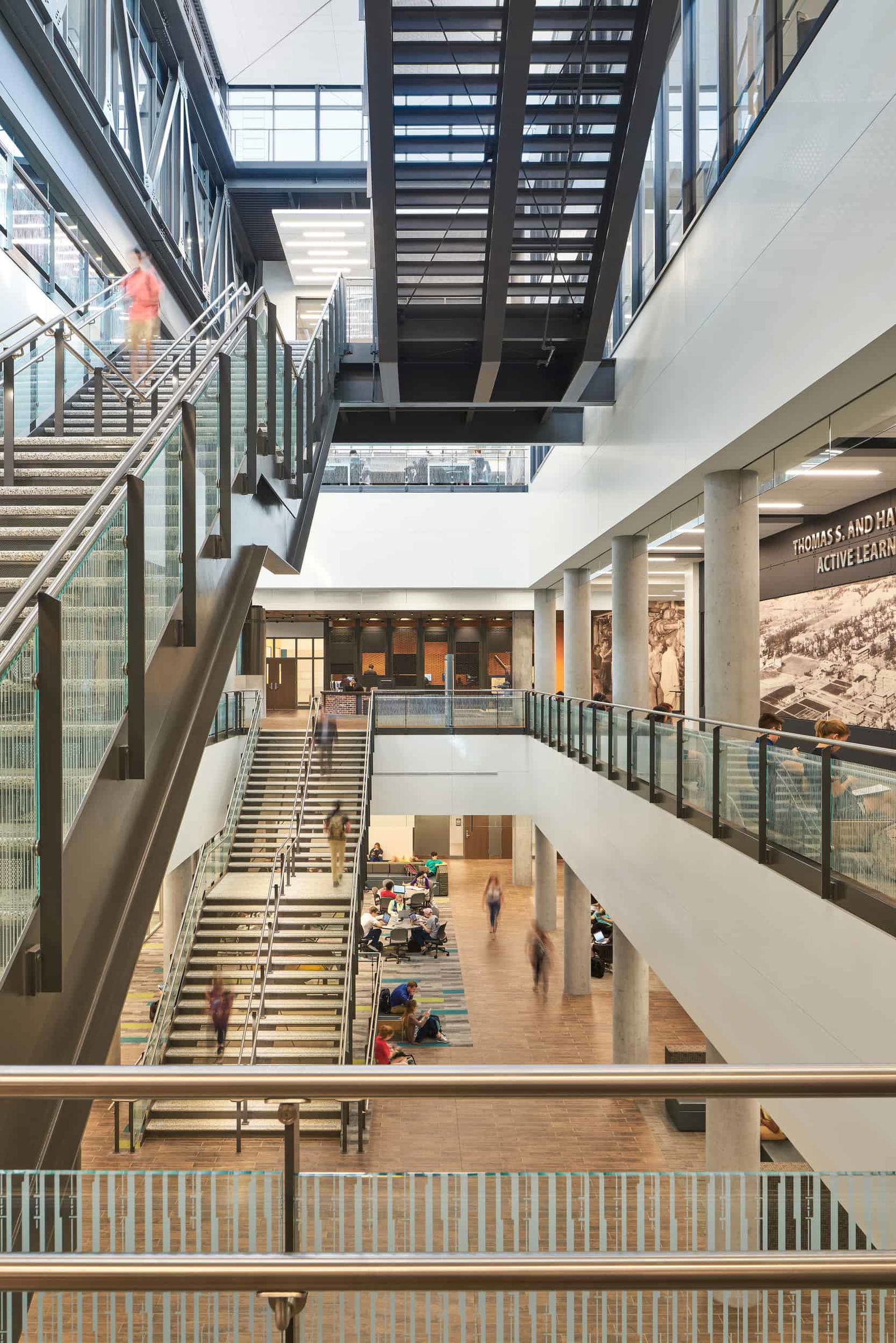 A spacious multi-level atrium in the Active Learning Center at Purdue University. The central feature is a large staircase with glass railings leading to multiple floors, with people ascending and descending. An open seating area on the ground floor has students gathered, some sitting on the floor while others work at tables. The space is illuminated by natural light streaming in through large windows. Metal walkways and staircases create a contemporary industrial aesthetic, complemented by concrete columns and glass partitions. A large mural or artwork is visible on the right wall, adding visual interest to the space.
