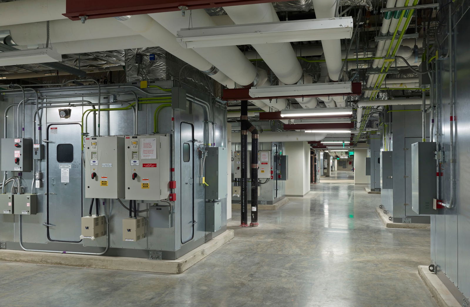 A well-lit industrial utility corridor inside the Sidney & Lois Eskenazi Hospital, featuring large HVAC and electrical systems. The space includes exposed pipes, ducts, and electrical panels mounted on metallic walls, with overhead fluorescent lighting illuminating the polished concrete floor. This image showcases BSA's expertise in engineering systems.