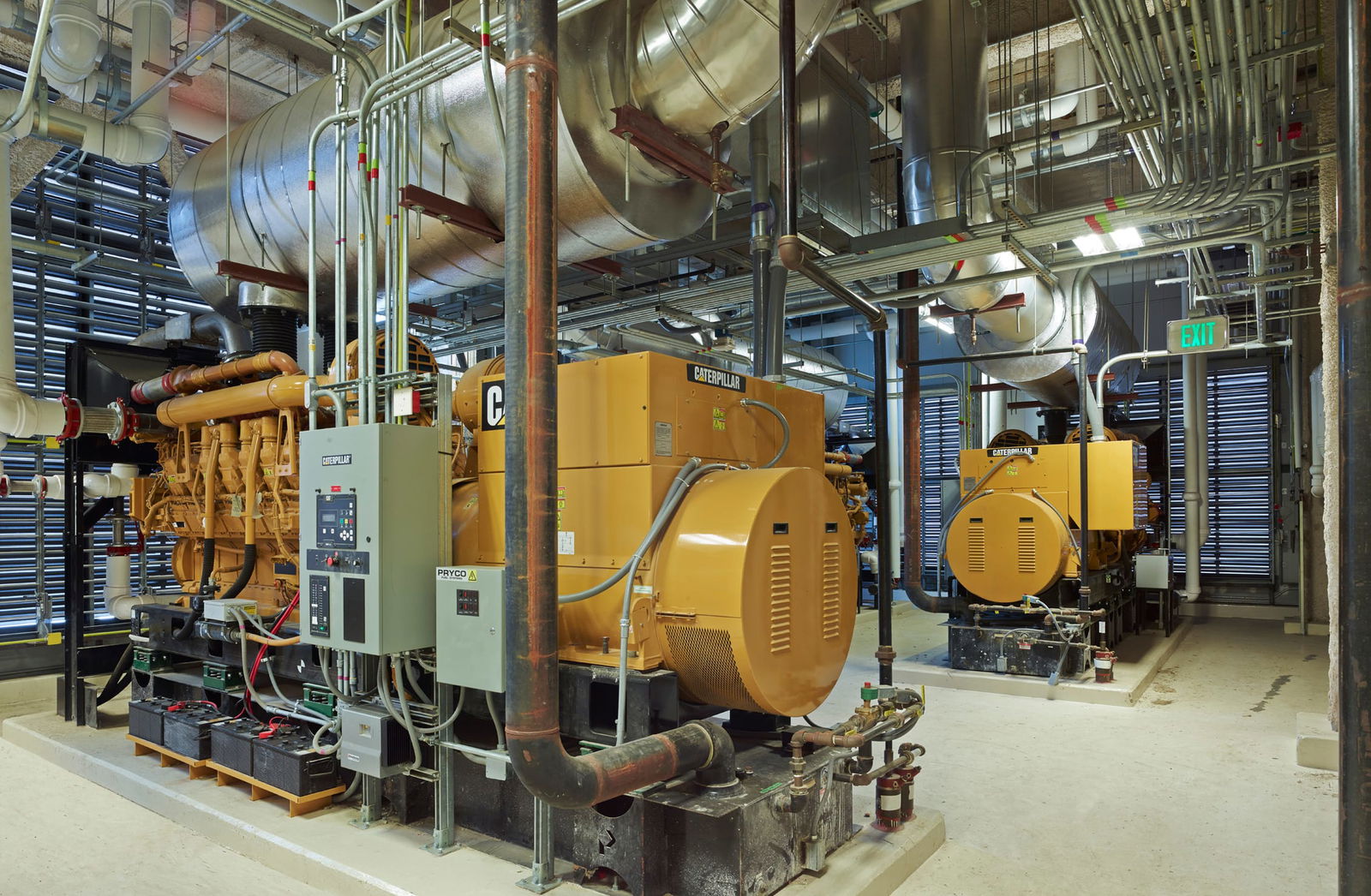 A well-lit industrial utility corridor inside the Sidney & Lois Eskenazi Hospital, featuring large HVAC and electrical systems. The space includes exposed pipes, ducts, and electrical panels mounted on metallic walls, with overhead fluorescent lighting illuminating the polished concrete floor. This image showcases BSA's expertise in MEP engineering systems.
