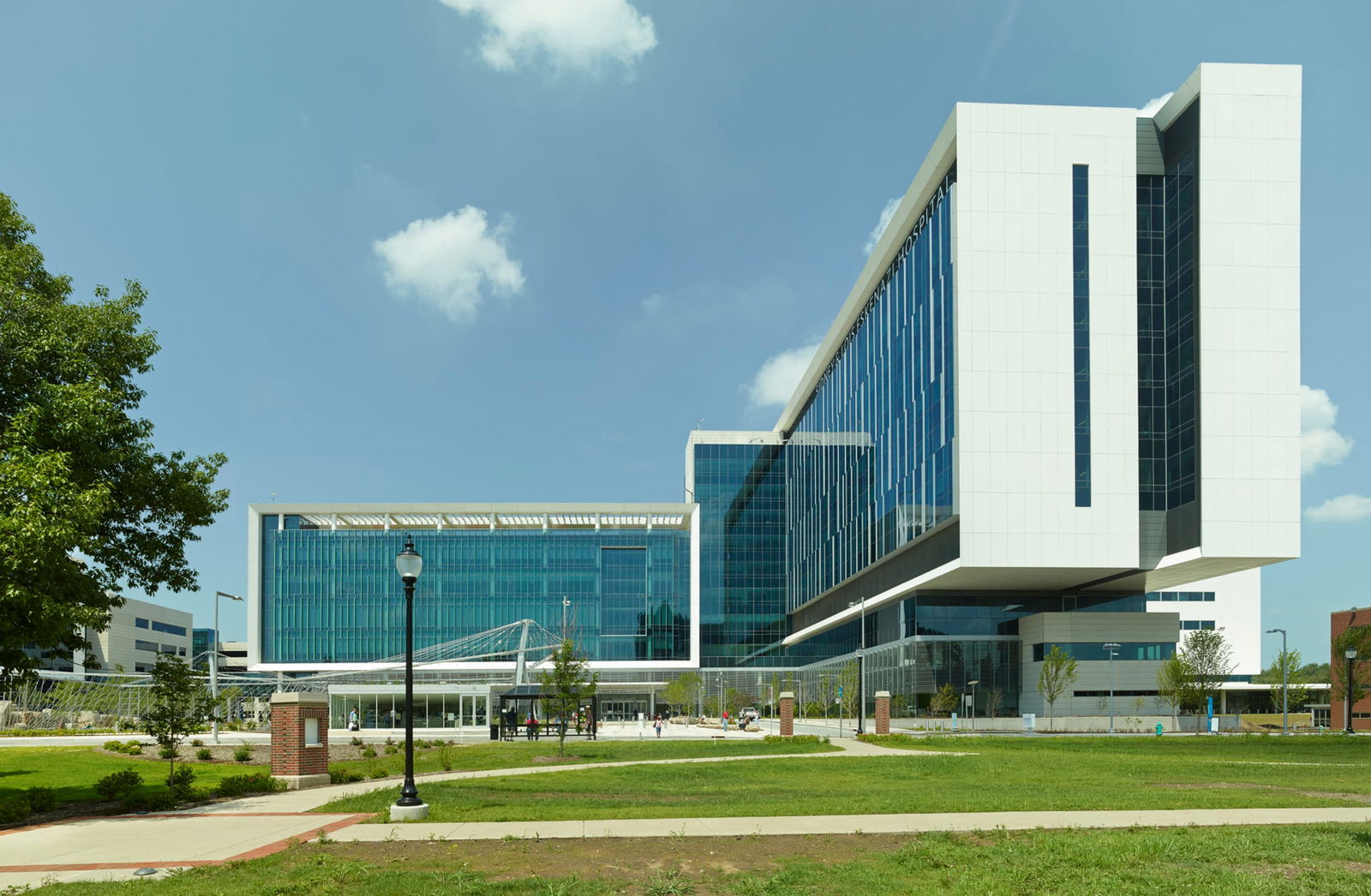 A modern hospital building with a sleek glass facade and white paneling, part of the Sidney & Lois Eskenazi Hospital. The structure features an elevated section extending outward, with a landscaped green space and walking paths in the foreground. The sky is clear with a few clouds, and pedestrians are visible near the entrance. This image showcases BSA's expertise in architecture.