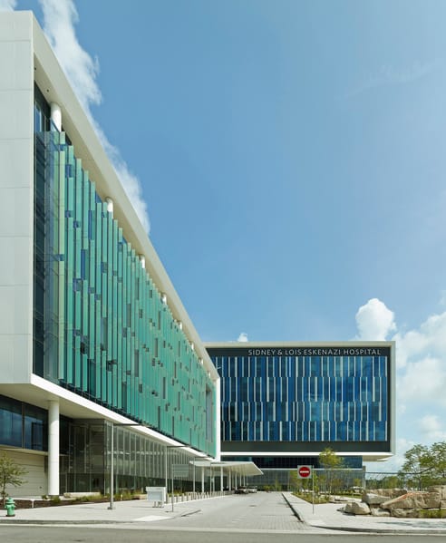 A modern hospital building with a sleek glass facade and white paneling, part of the Sidney & Lois Eskenazi Hospital. The structure features an elevated section extending outward, with a landscaped green space and walking paths in the foreground. The sky is clear with a few clouds, and pedestrians are visible near the entrance. This image showcases BSA's expertise in architecture.
