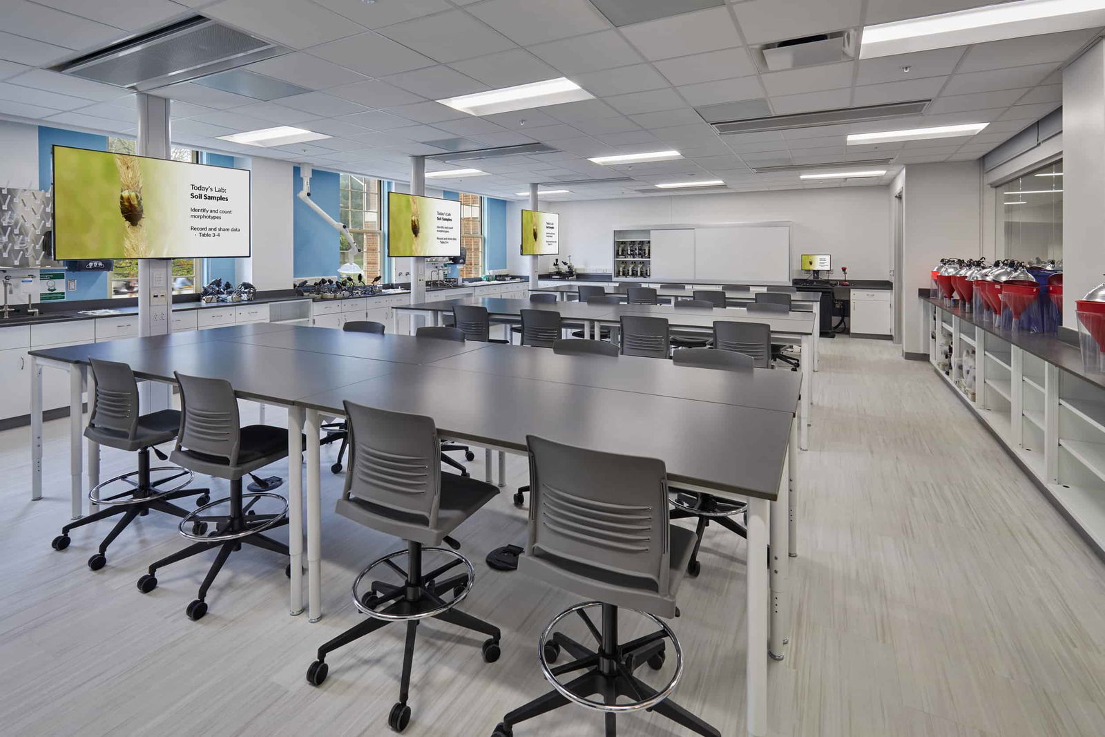 Bright, efficient, and future-focused, this teaching lab is designed to support hands-on experiential learning. The modular lab benches and mobile stools accommodate flexible group sizes and encourage interaction. Dual high-definition monitors provide instructional content to all corners of the room, minimizing obstruction and enhancing clarity. Along the walls, microscopes, experimental stations, and other equipment stand ready for biological analysis—in this case, a lab on soil samples and insect identification. Natural light filters through large windows, and the space is cleanly organized with ample storage, reinforcing an environment of both precision and curiosity.
